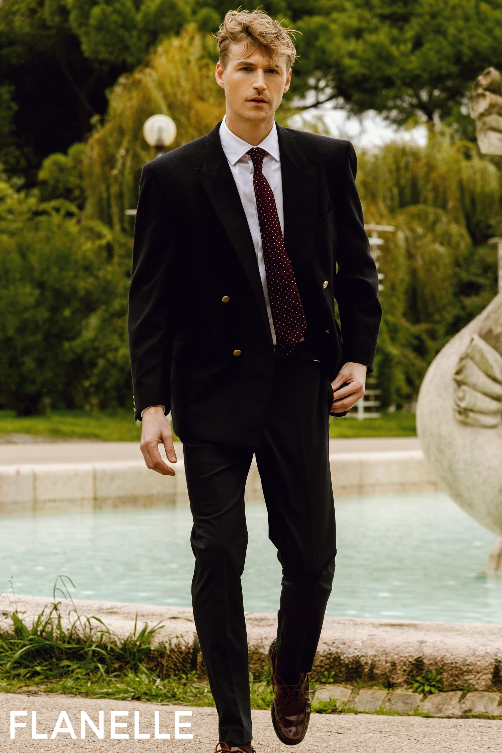 Man in stylish suit walking by a fountain in a garden setting.