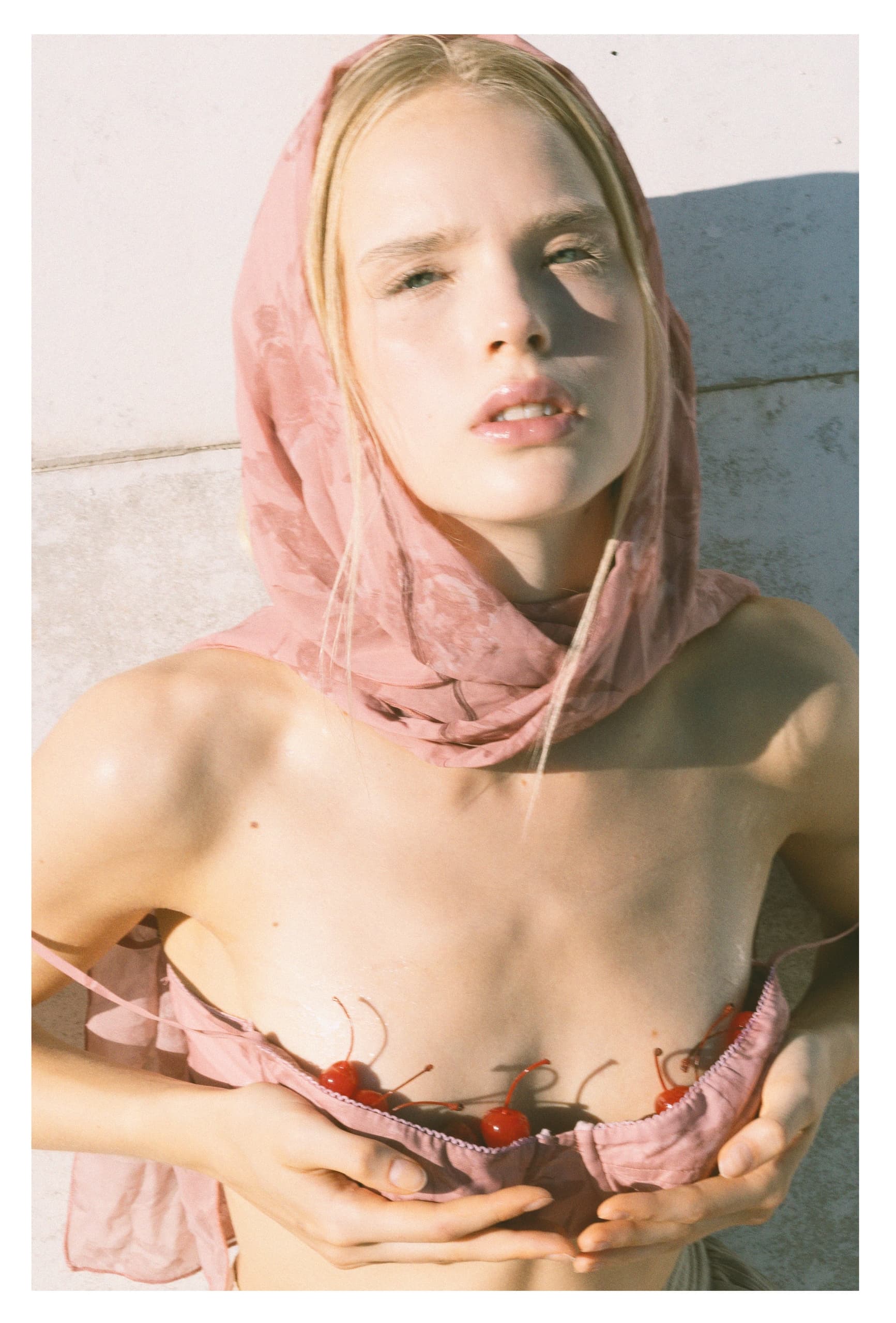 Young woman in pink scarf, holding cherries in pink fabric, sunlight on face.