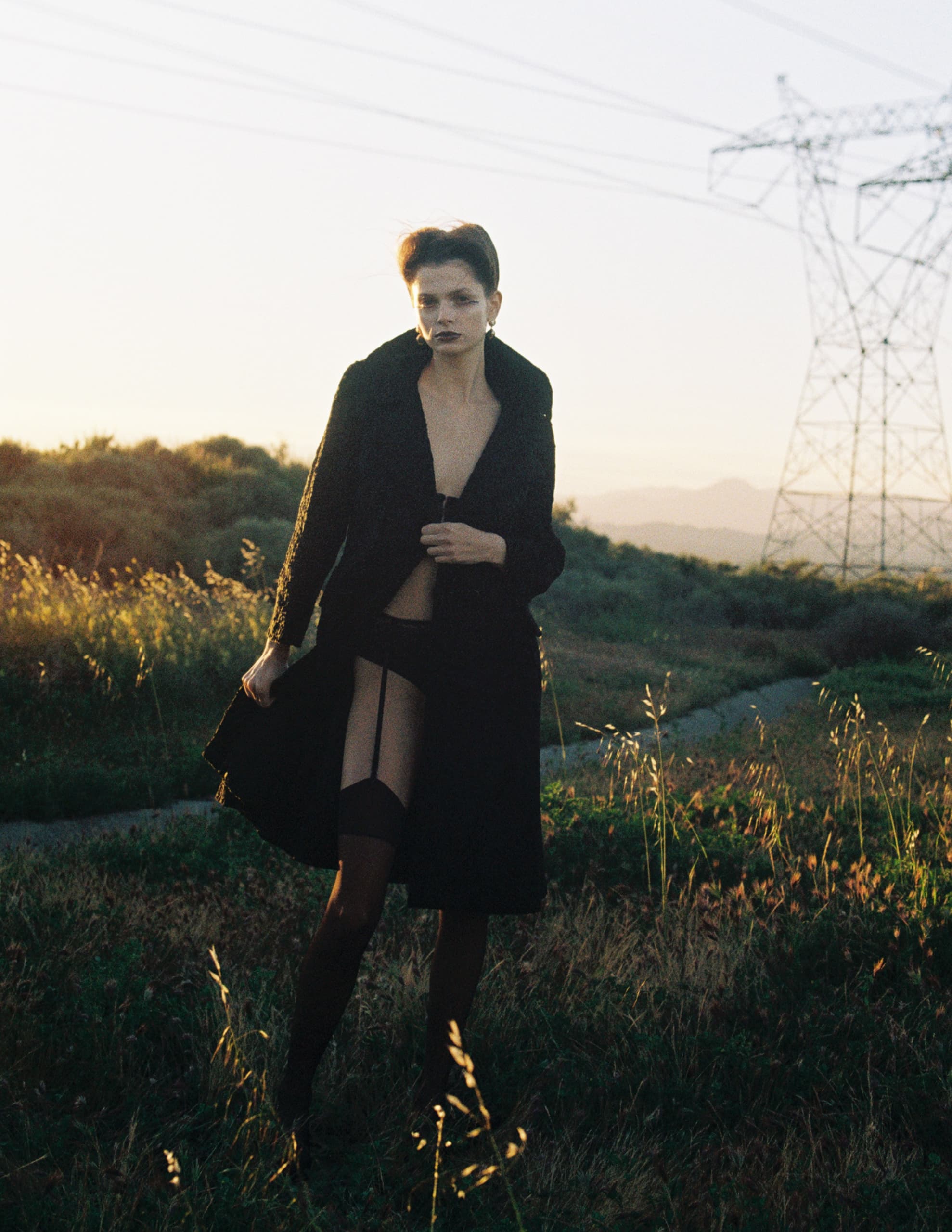 Woman in dark fashion poses outdoors near power lines at sunset.