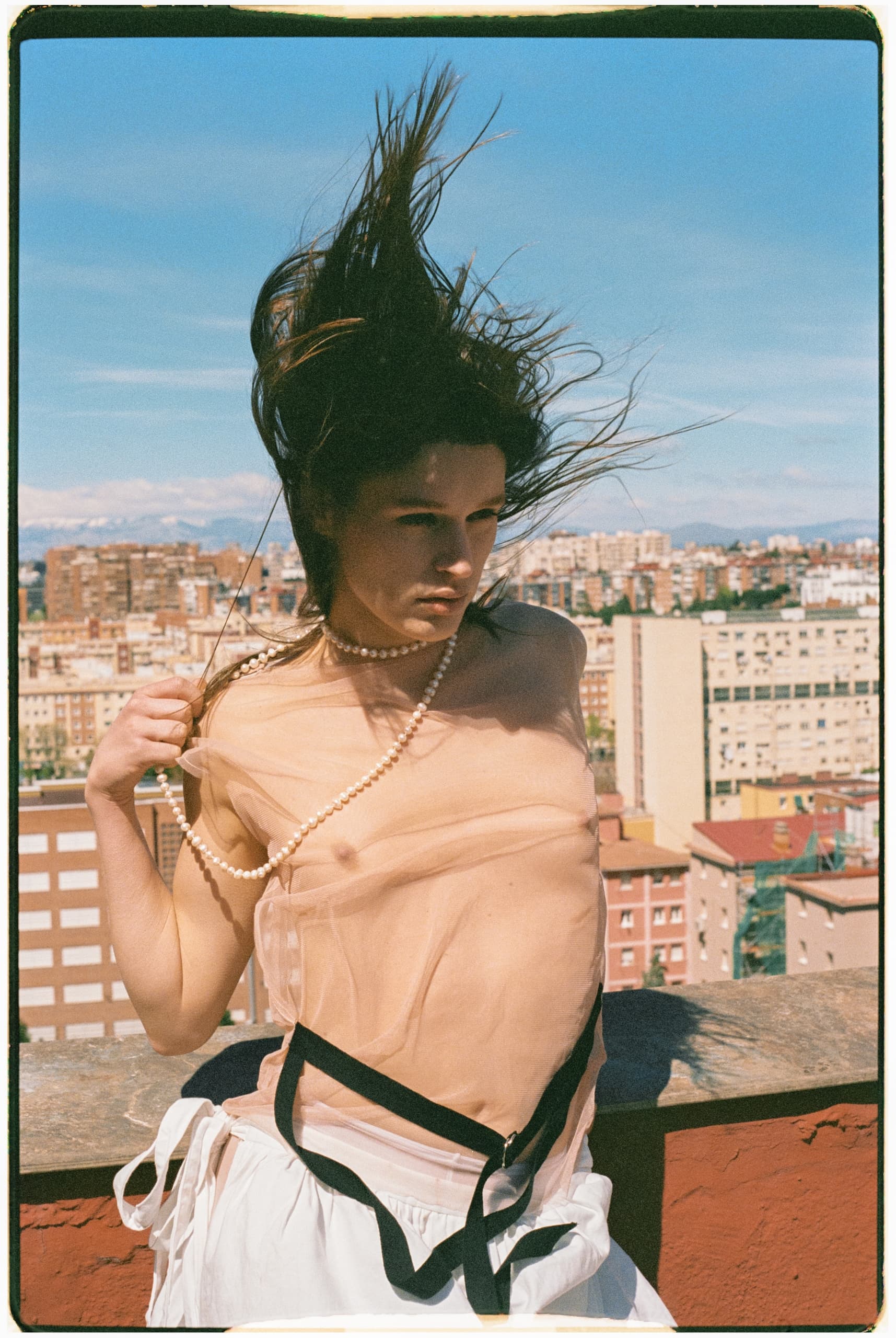 Model with flowing hair and pearl necklace on rooftop with city skyline background.