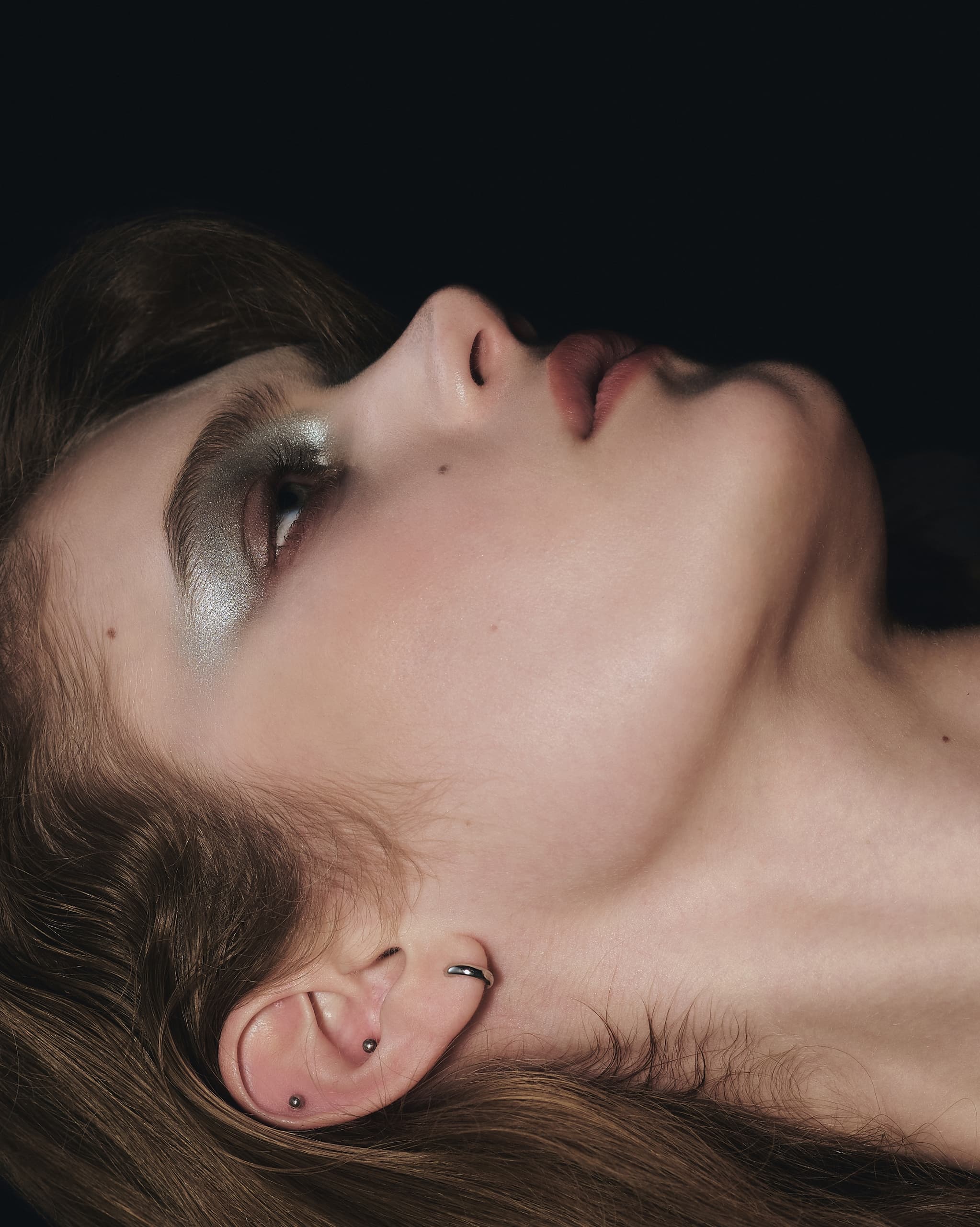 Close-up of a woman with silver eyeshadow and ear piercings, tilted head.