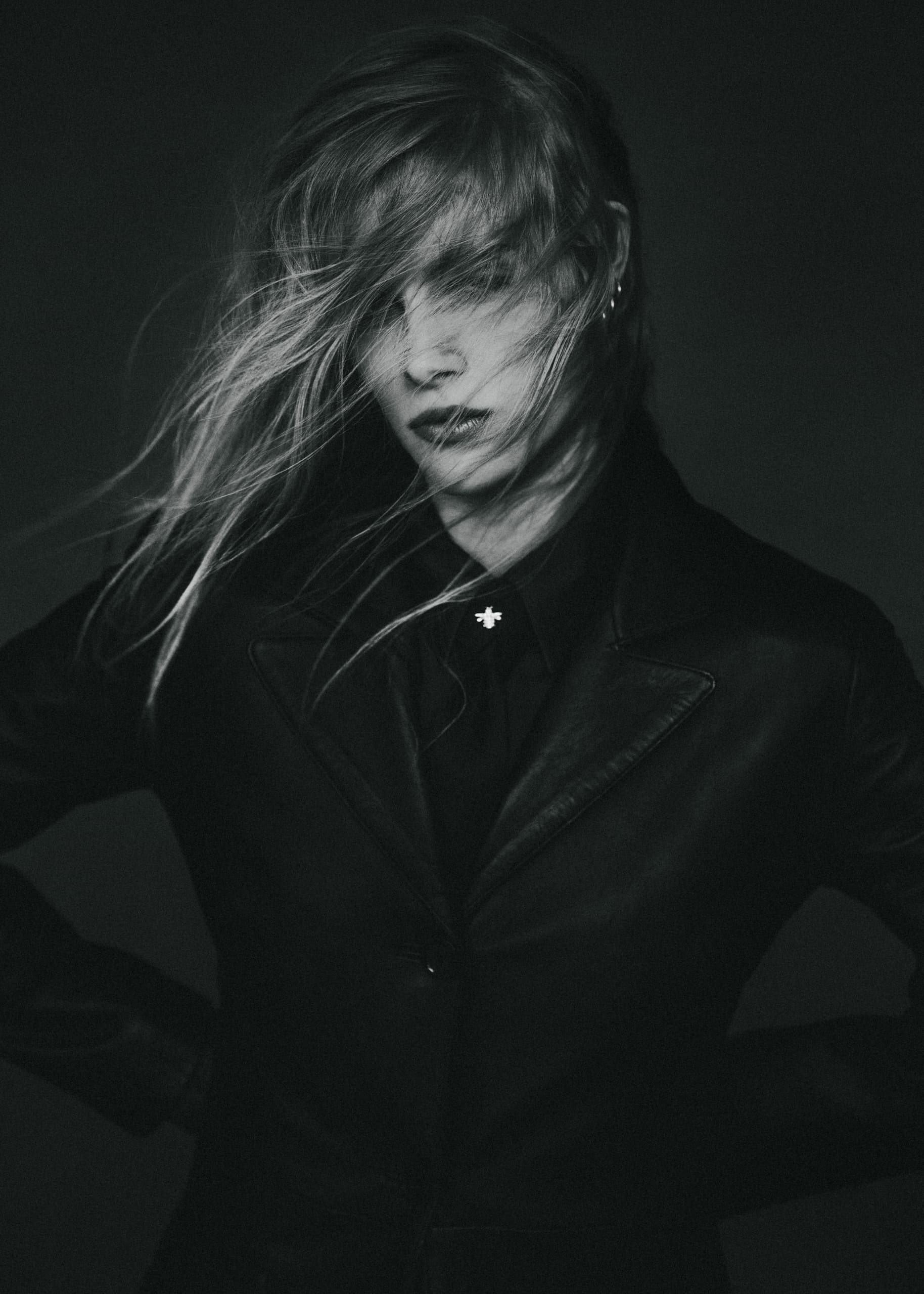 Mysterious woman in black coat, hair windswept, moody black-and-white portrait.