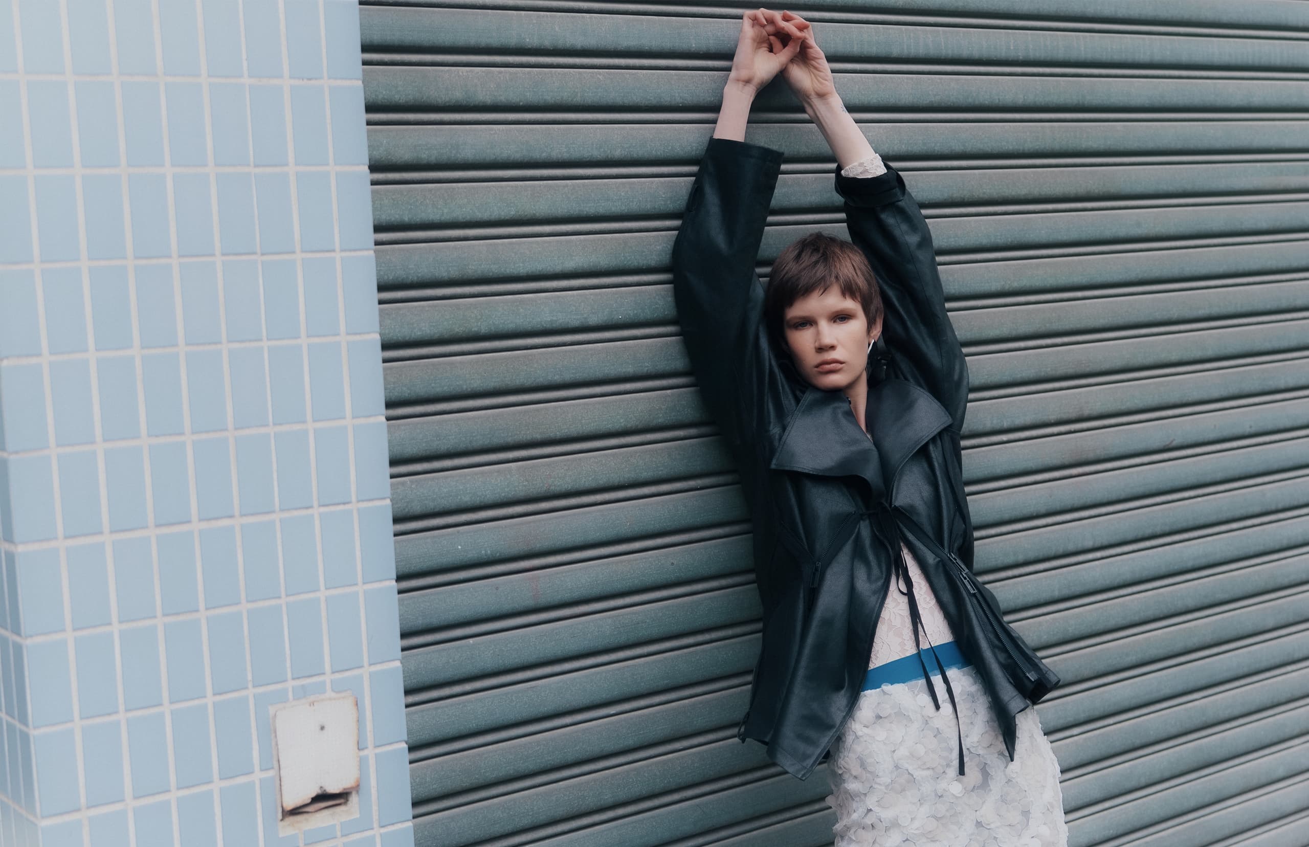 Fashionable person posing against blue tiled wall and metal shutter, wearing black jacket.