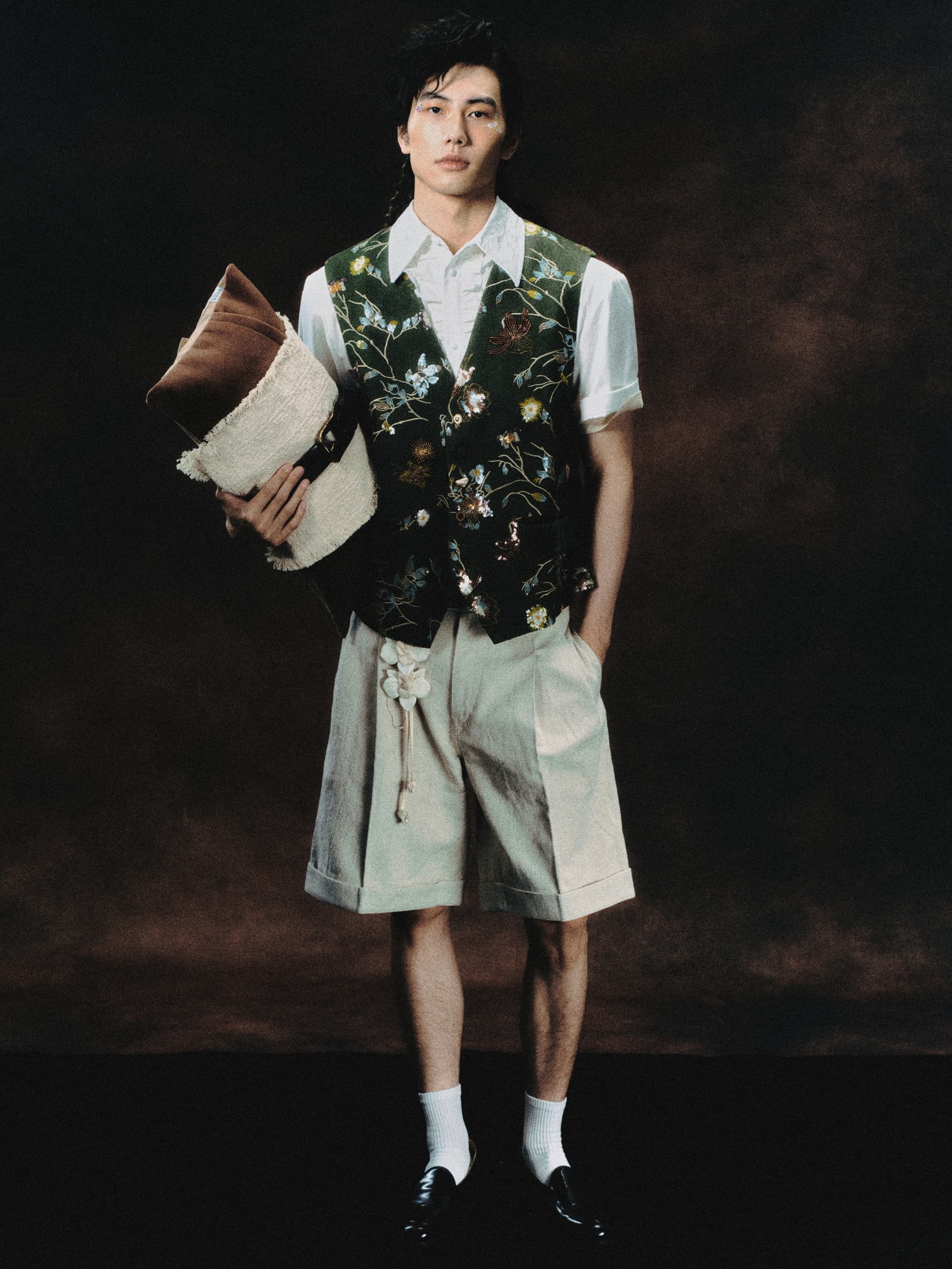 Man in floral vest and shorts holding pillows, standing against a dark background.