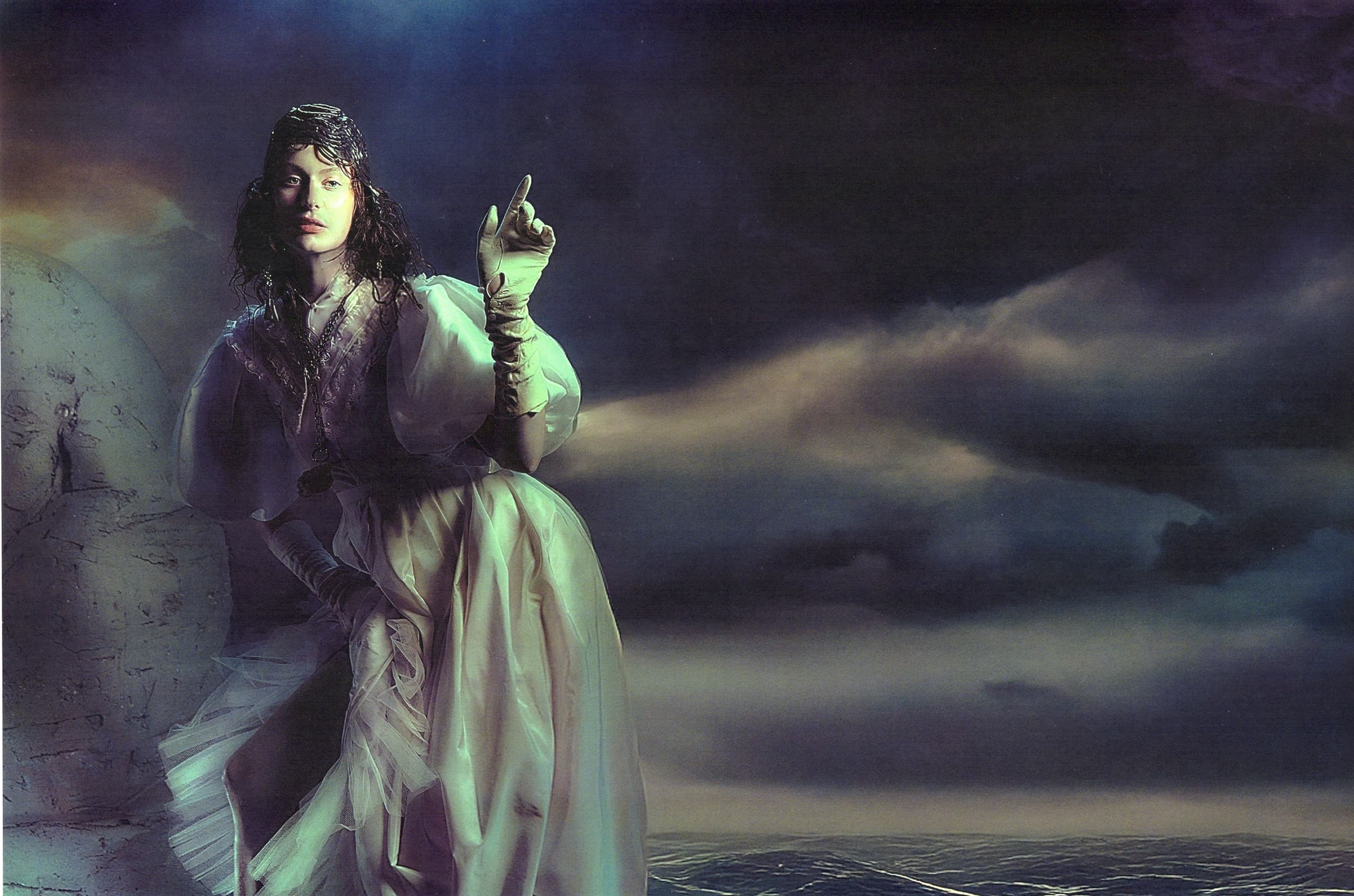 Mystical woman in white gown points upward amid dark stormy sea and sky.