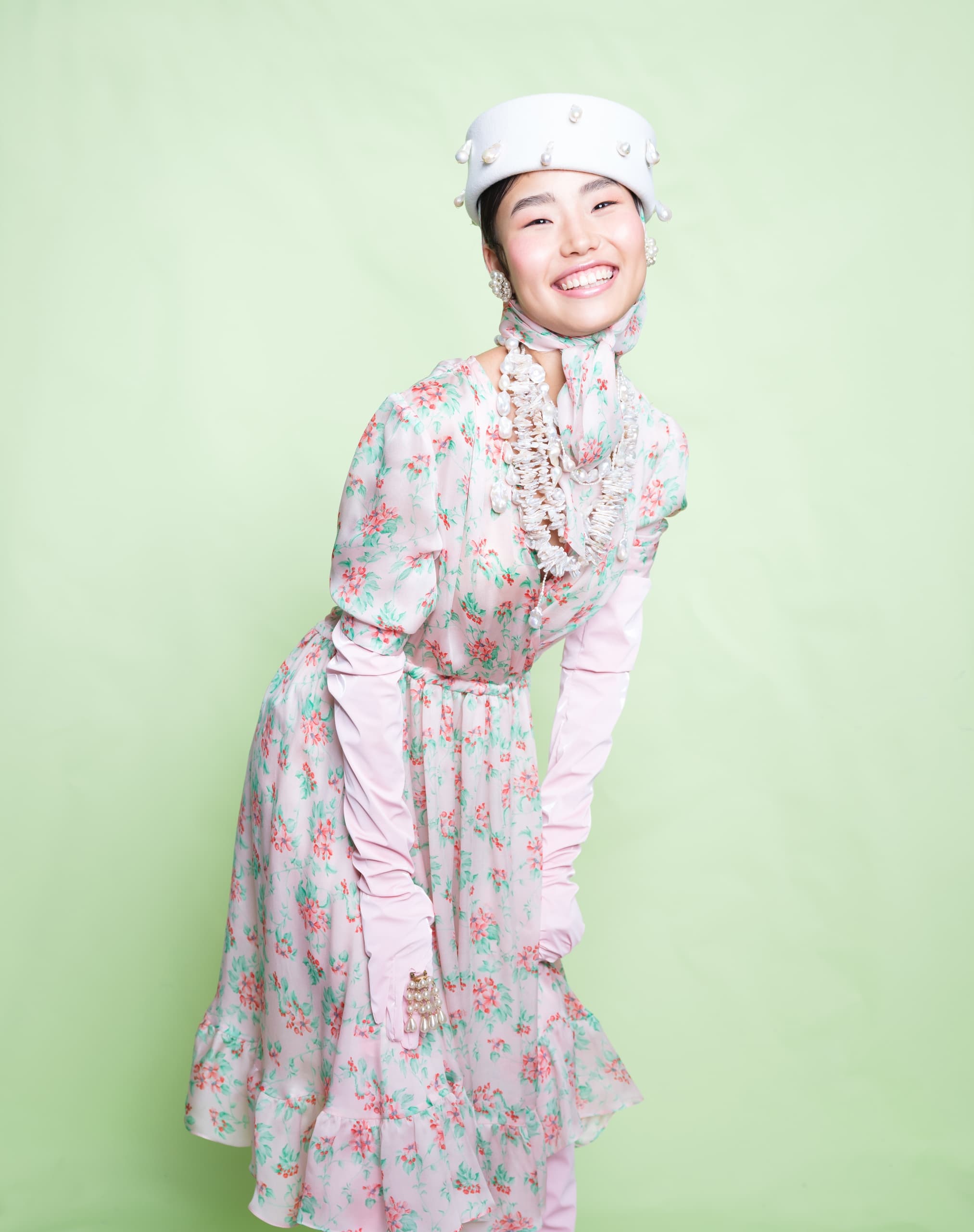 Smiling woman in floral dress and pearl hat on a light green background.