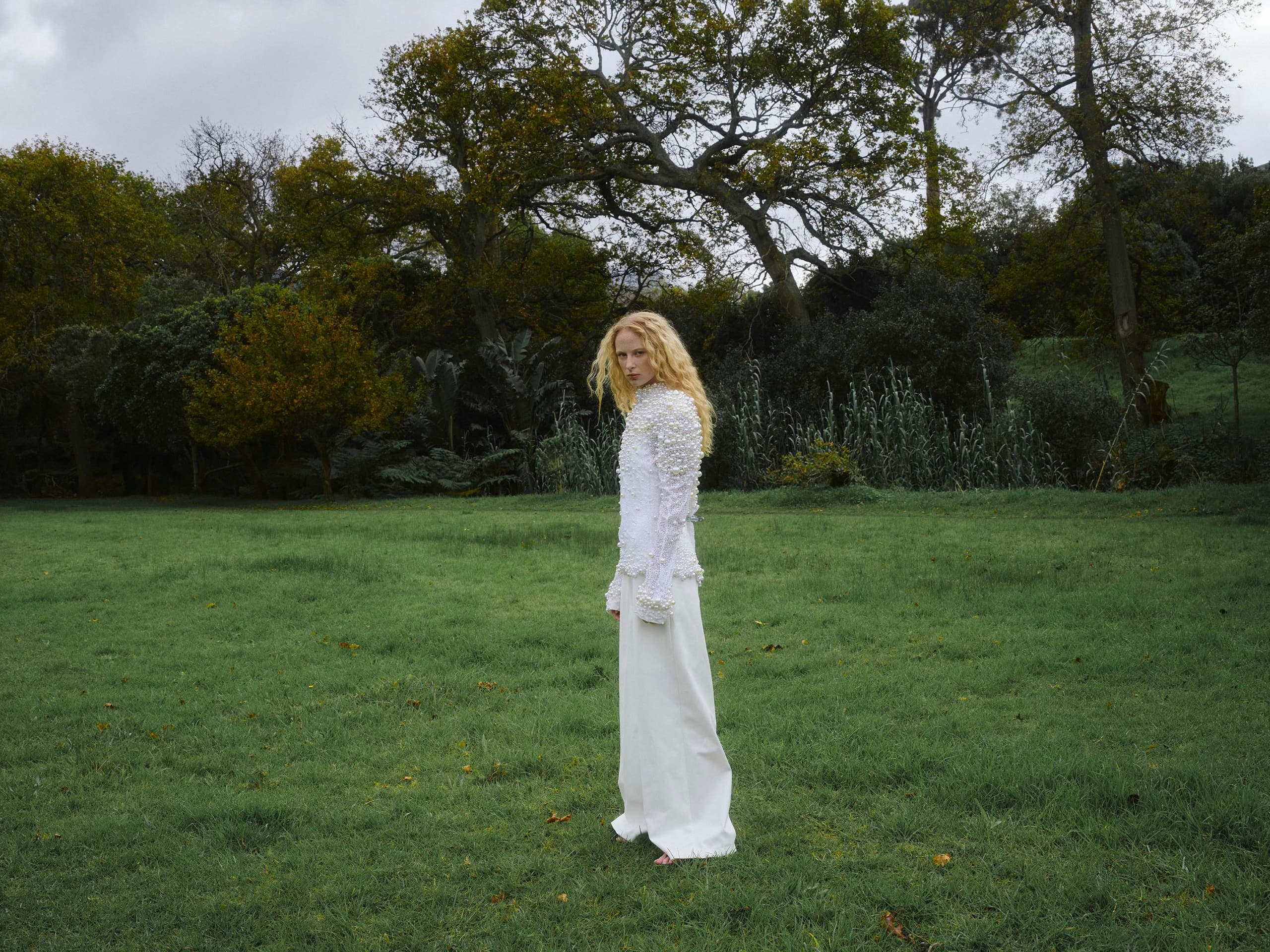 Person in white outfit standing in lush green park with trees in the background.