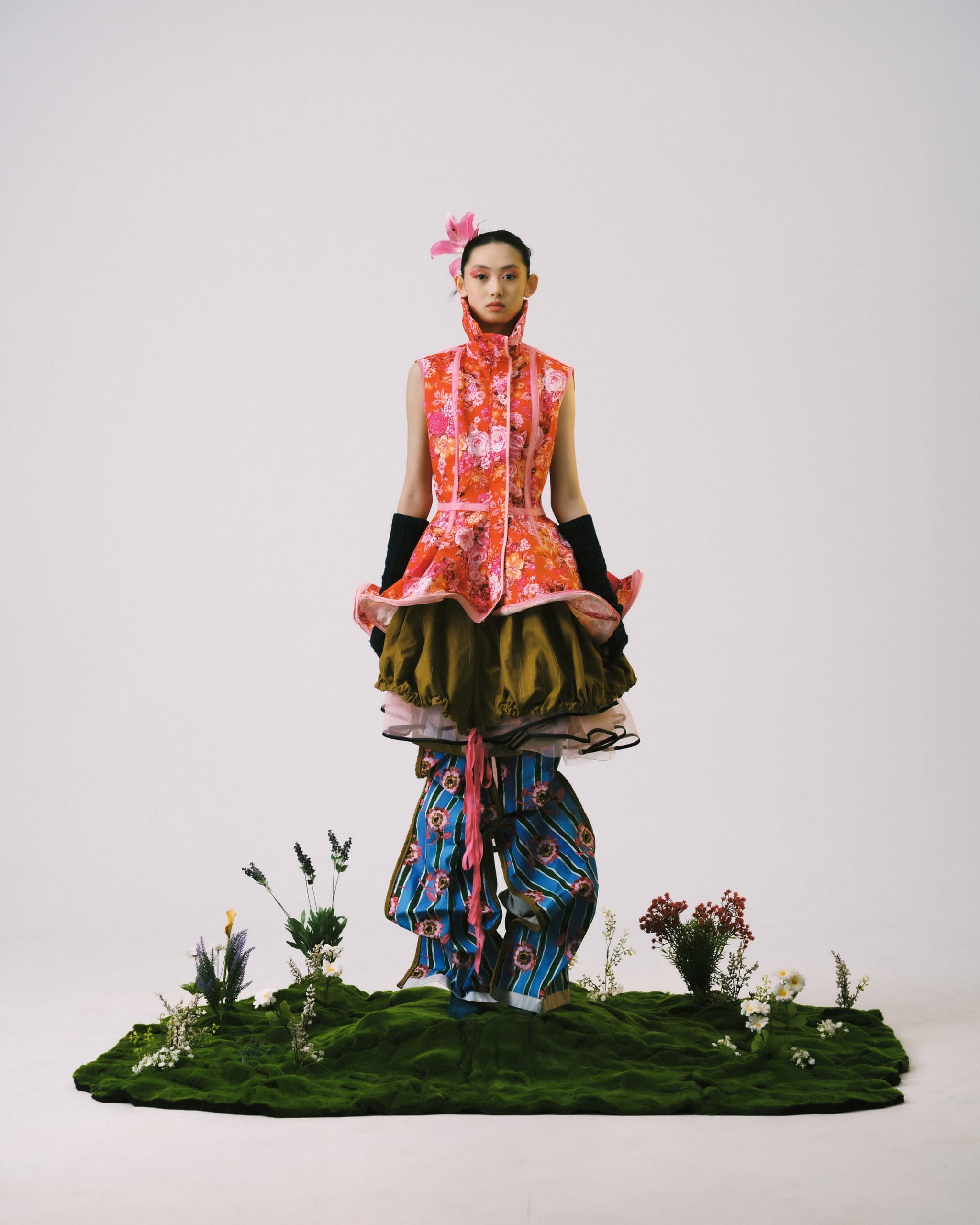 Avant-garde fashion model in colorful layered outfit on grass with flowers.