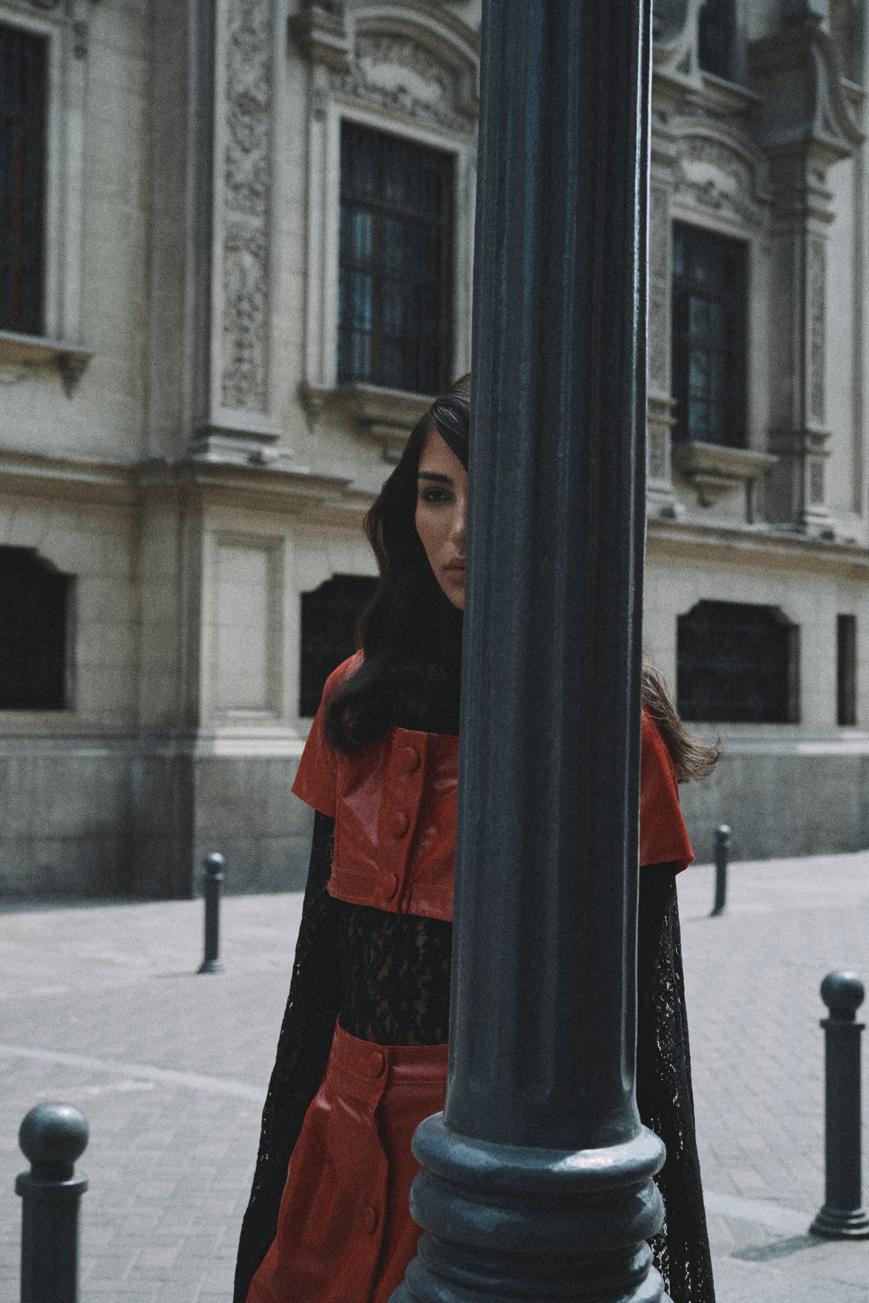 Woman half-hidden by pole in vintage urban setting, wearing red outfit.