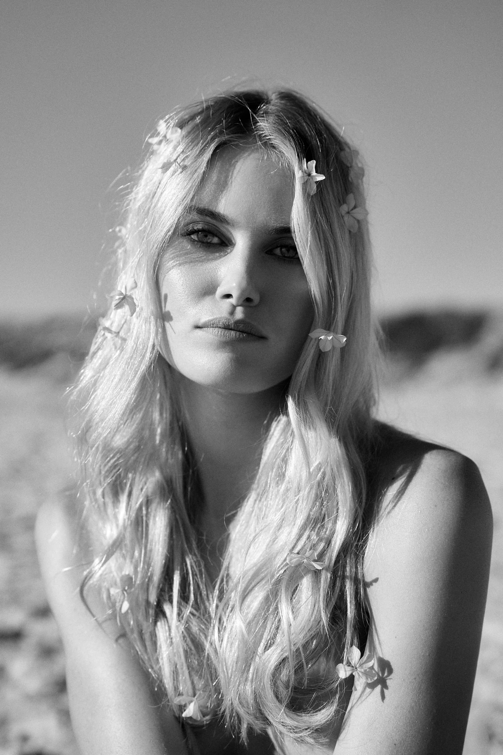 Black and white portrait of a woman with flowers in her hair, beach in background.