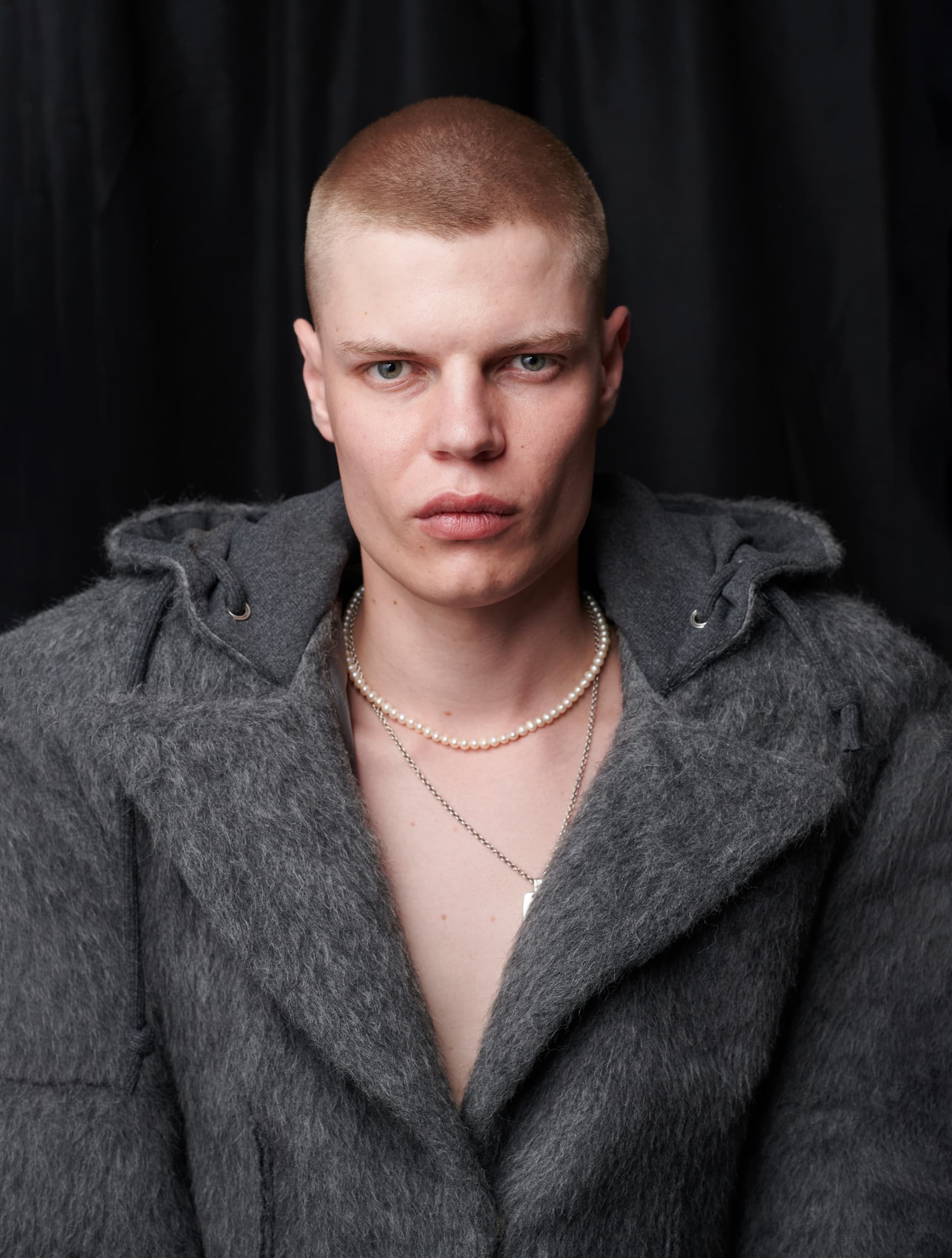 Trendy individual in gray coat with pearl necklaces against dark backdrop.