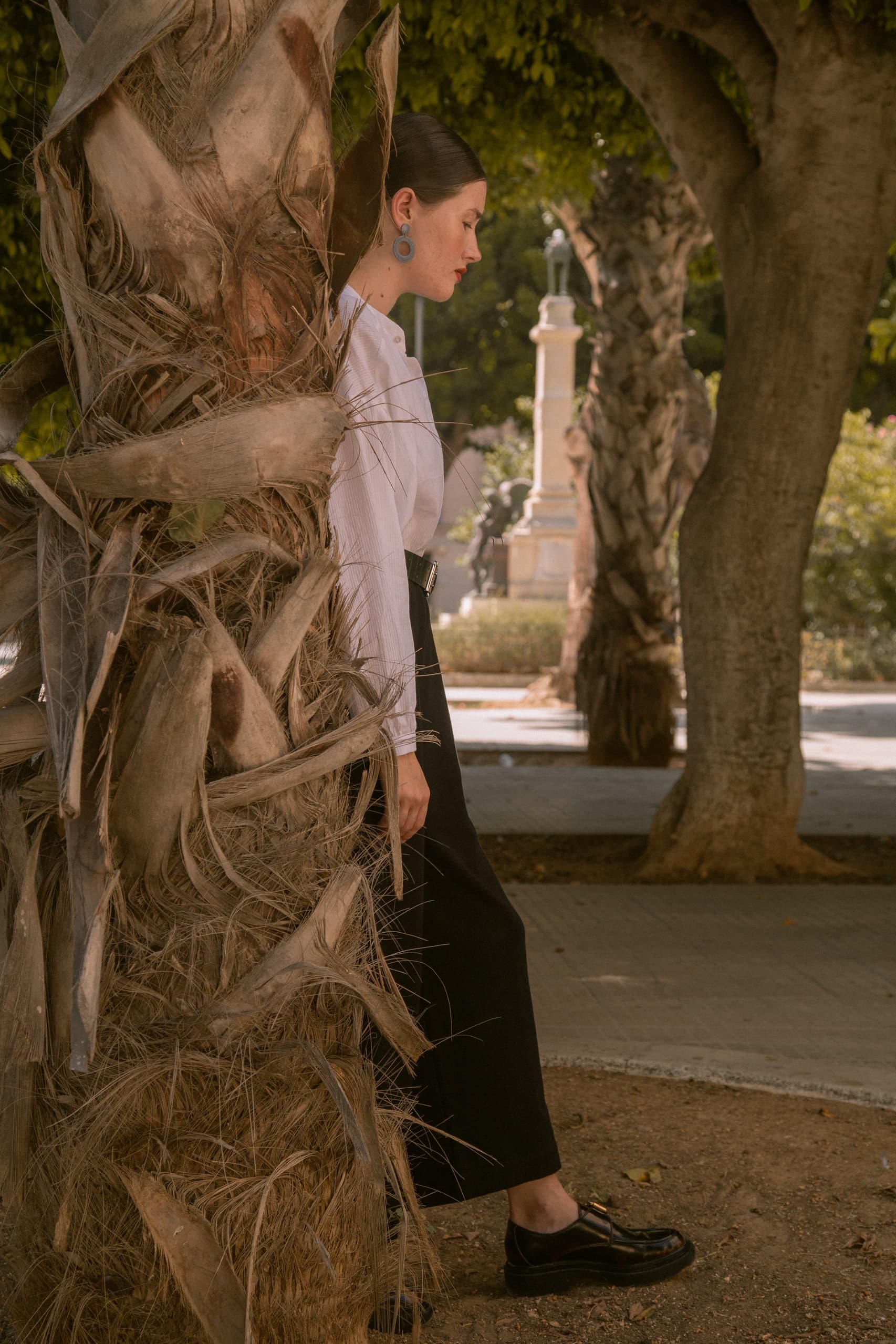 Person stands beside palm tree in a sunlit park, wearing a white blouse and dark pants.