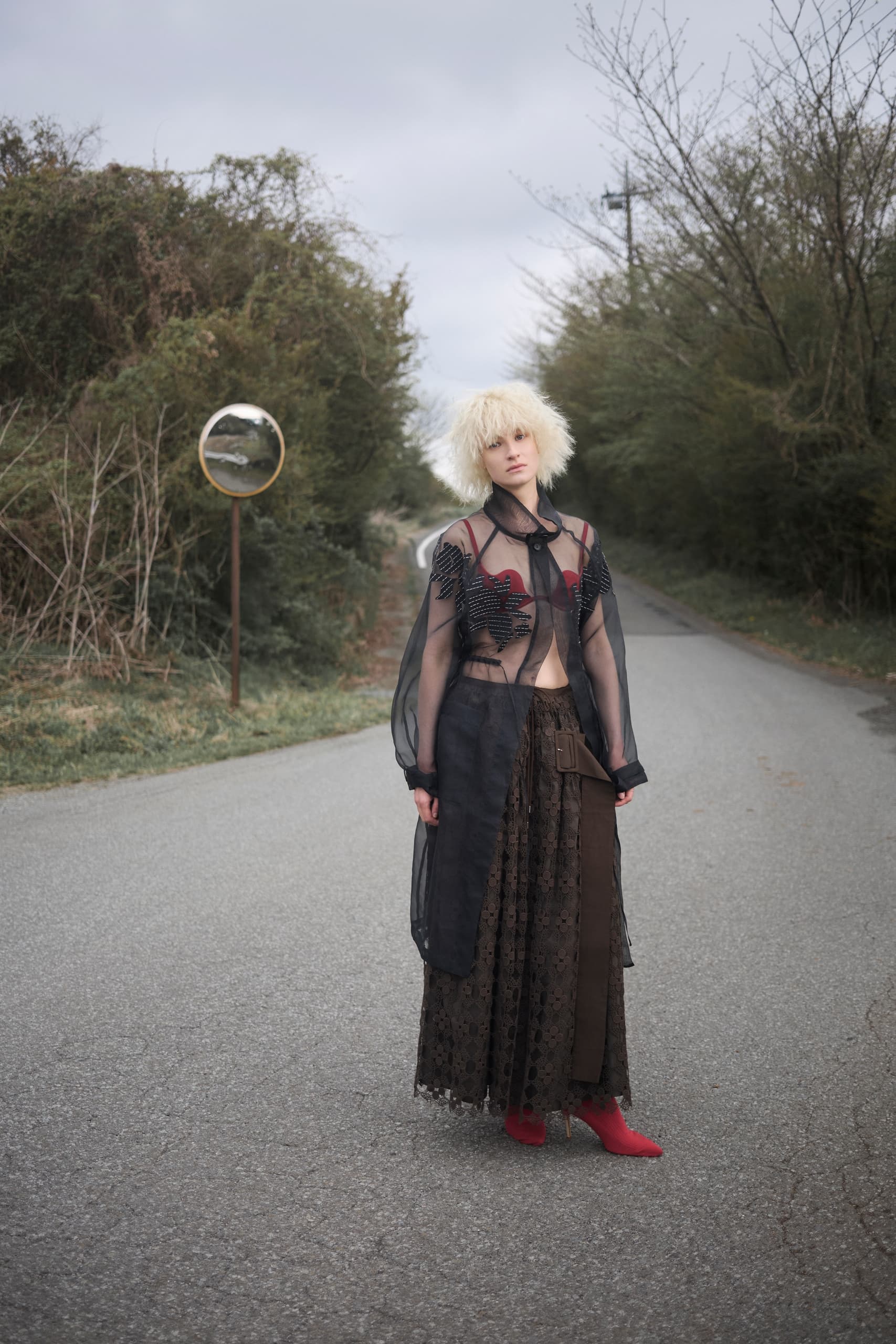 Fashionable woman in a sheer black dress stands on a rural road with trees.