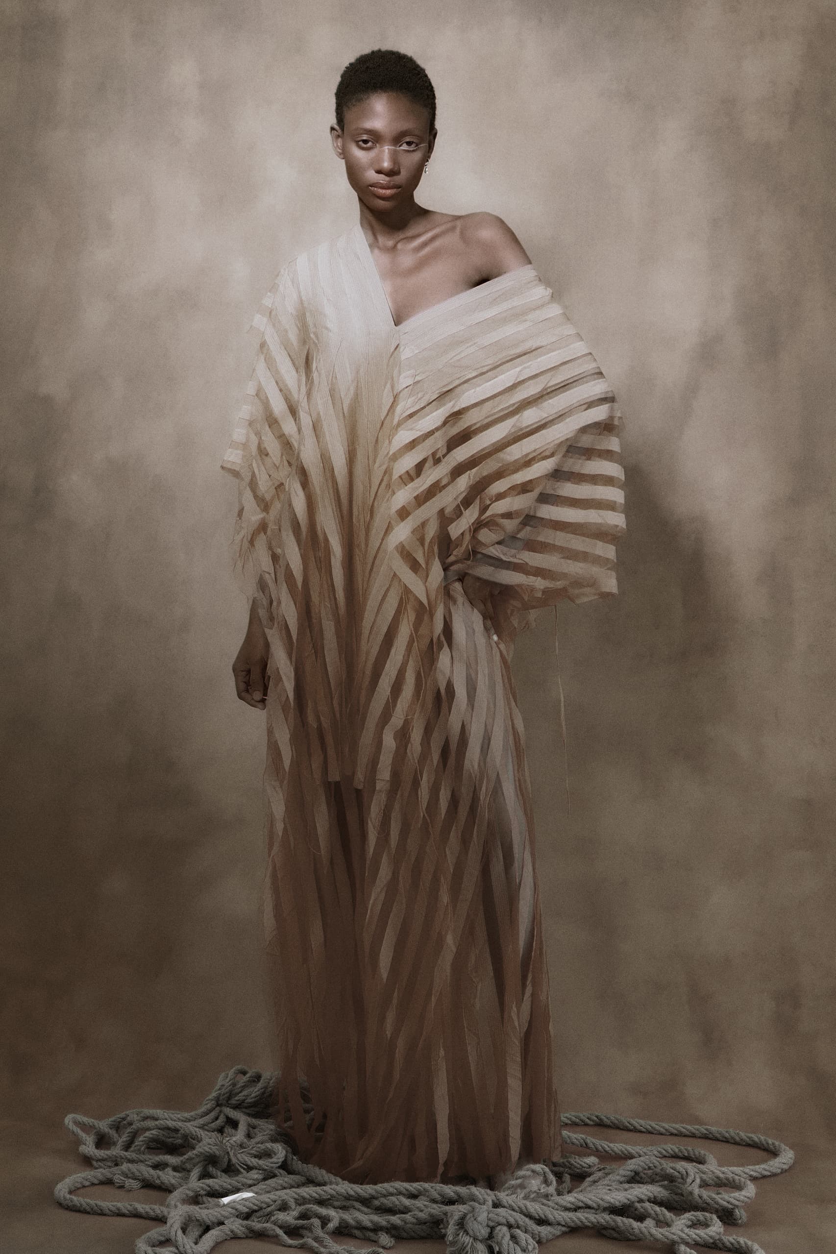 Model in a striped sheer gown standing on ropes against a beige backdrop.