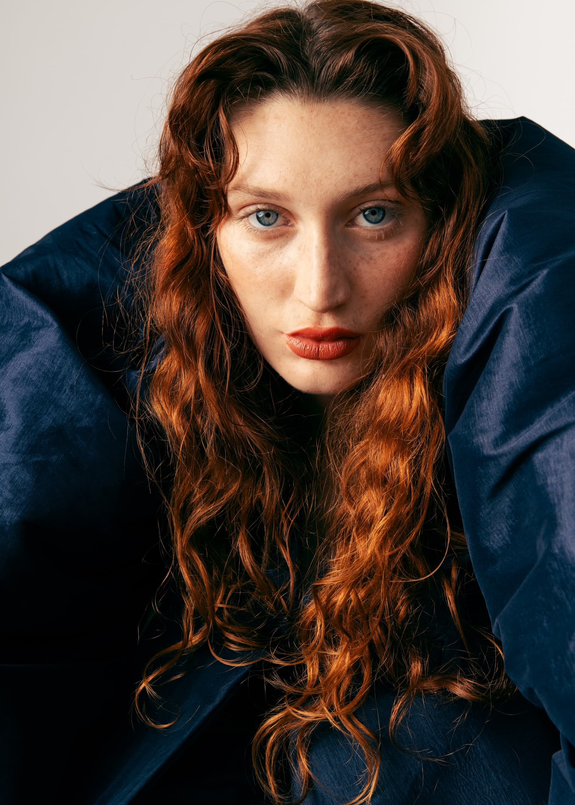 Woman with red wavy hair in a dark outfit, intense gaze, white background.