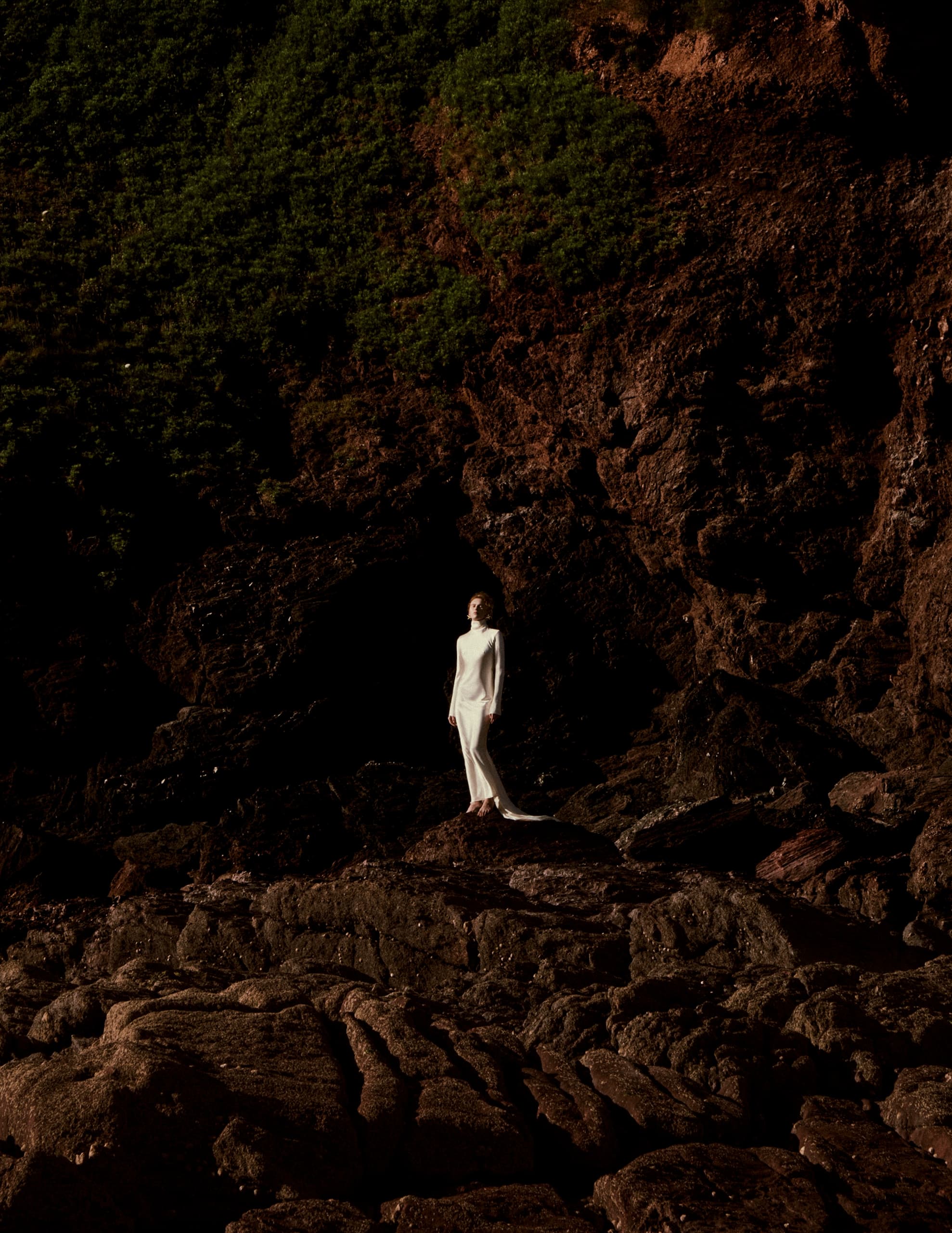 Person in white dress on rocky cliff with green foliage background.
