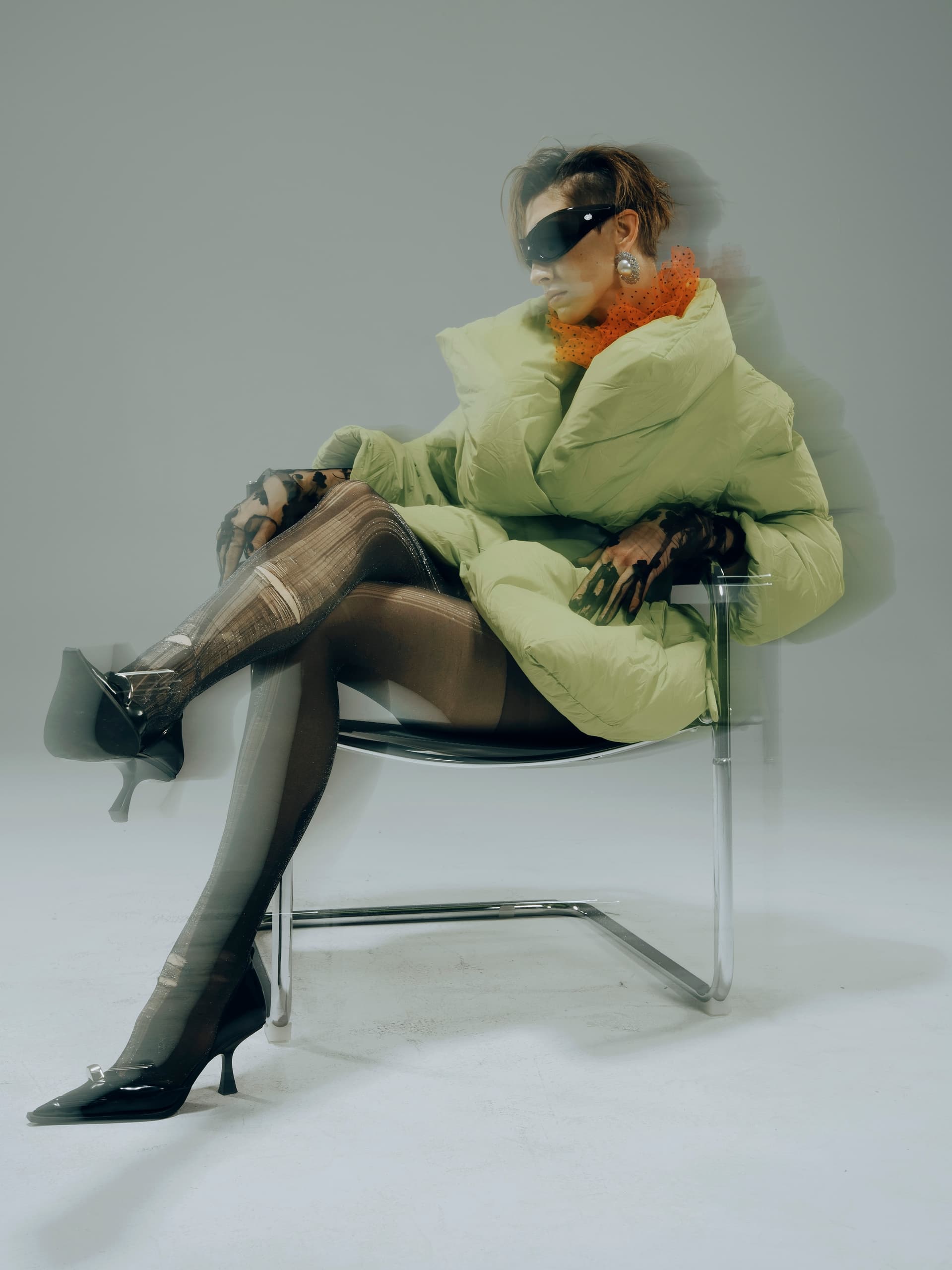 Futuristic fashion: woman in puffer coat and sunglasses on chair, artistic blur effect.