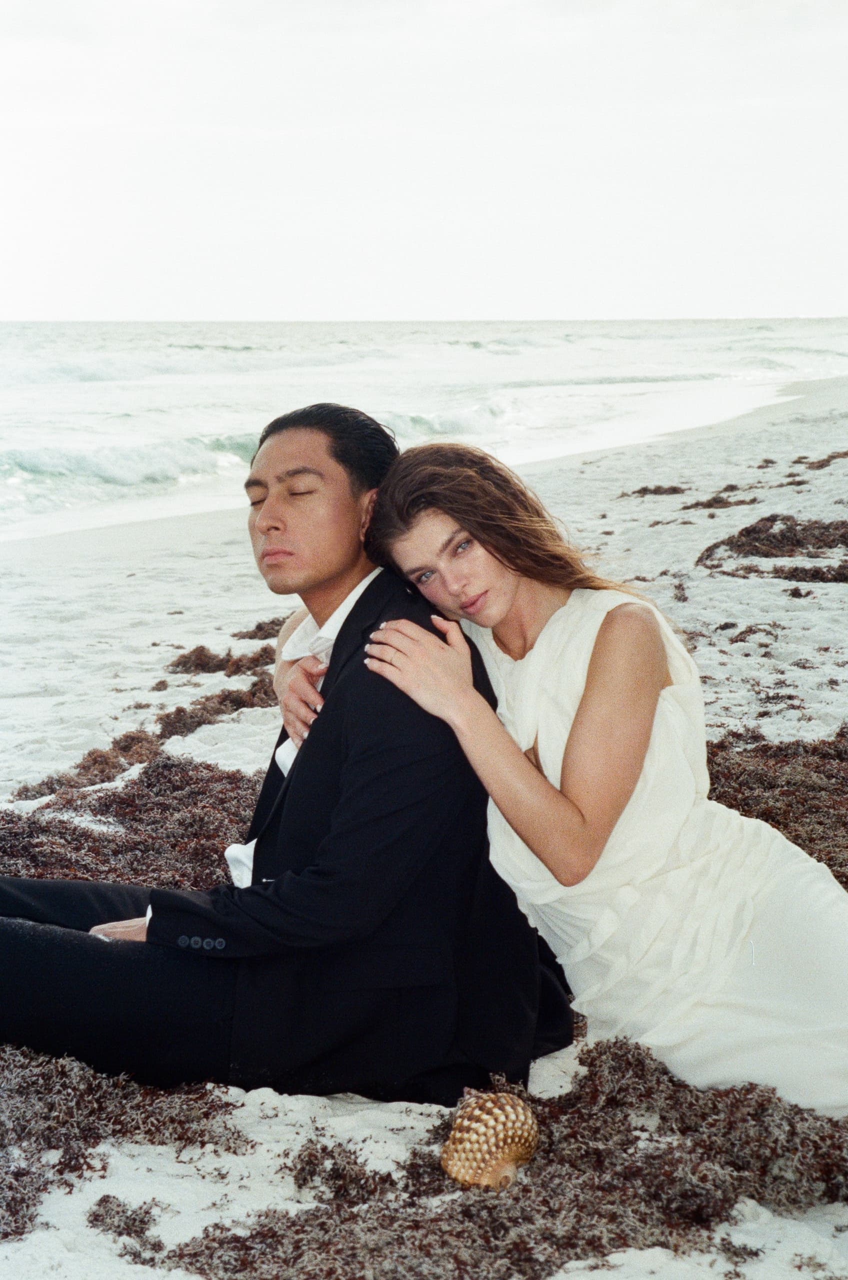 Couple in elegant attire embrace on a serene beach with ocean waves in the background.
