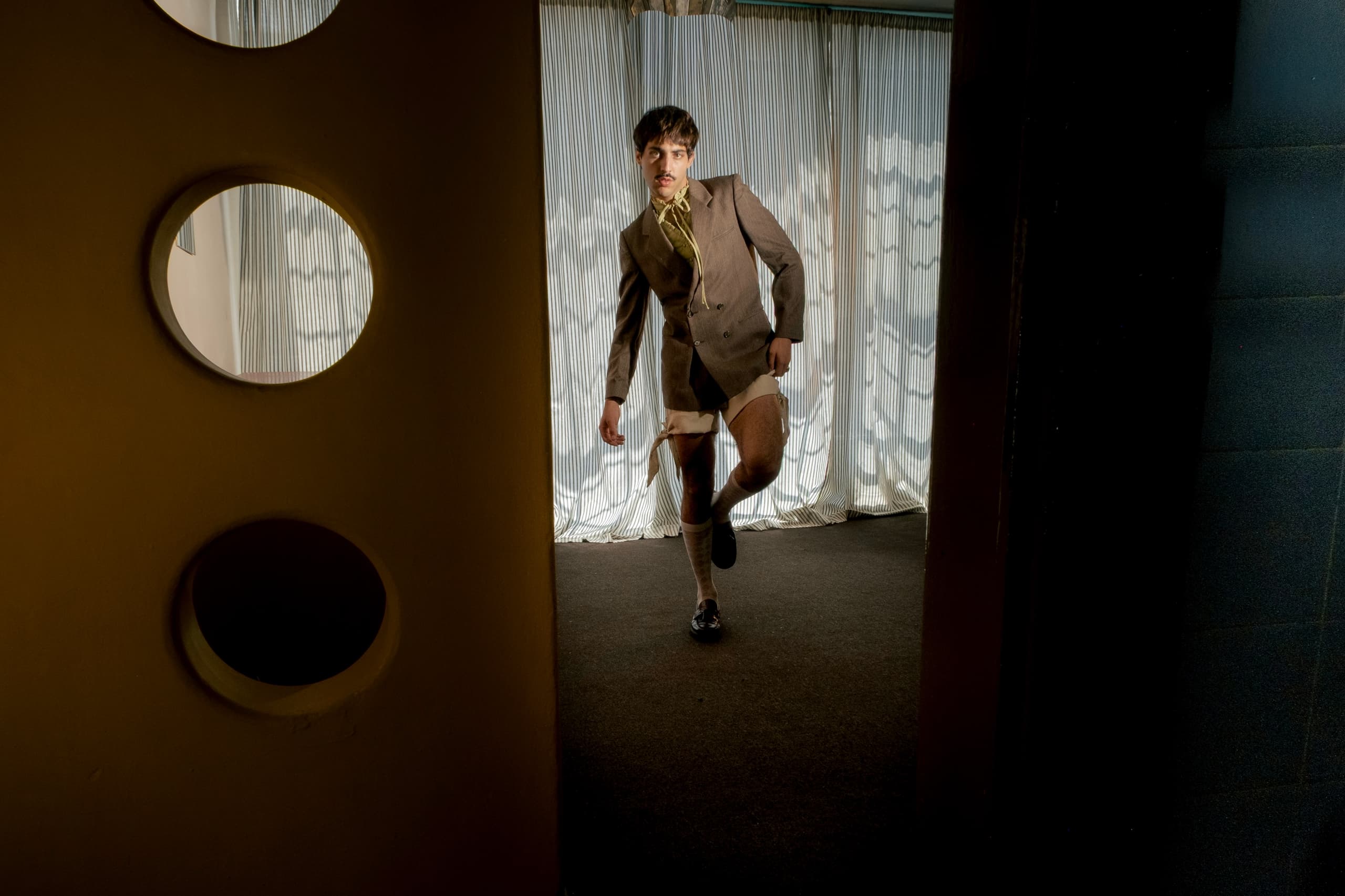 Person in a partially torn suit posing in a dimly lit room with curtain backdrop.