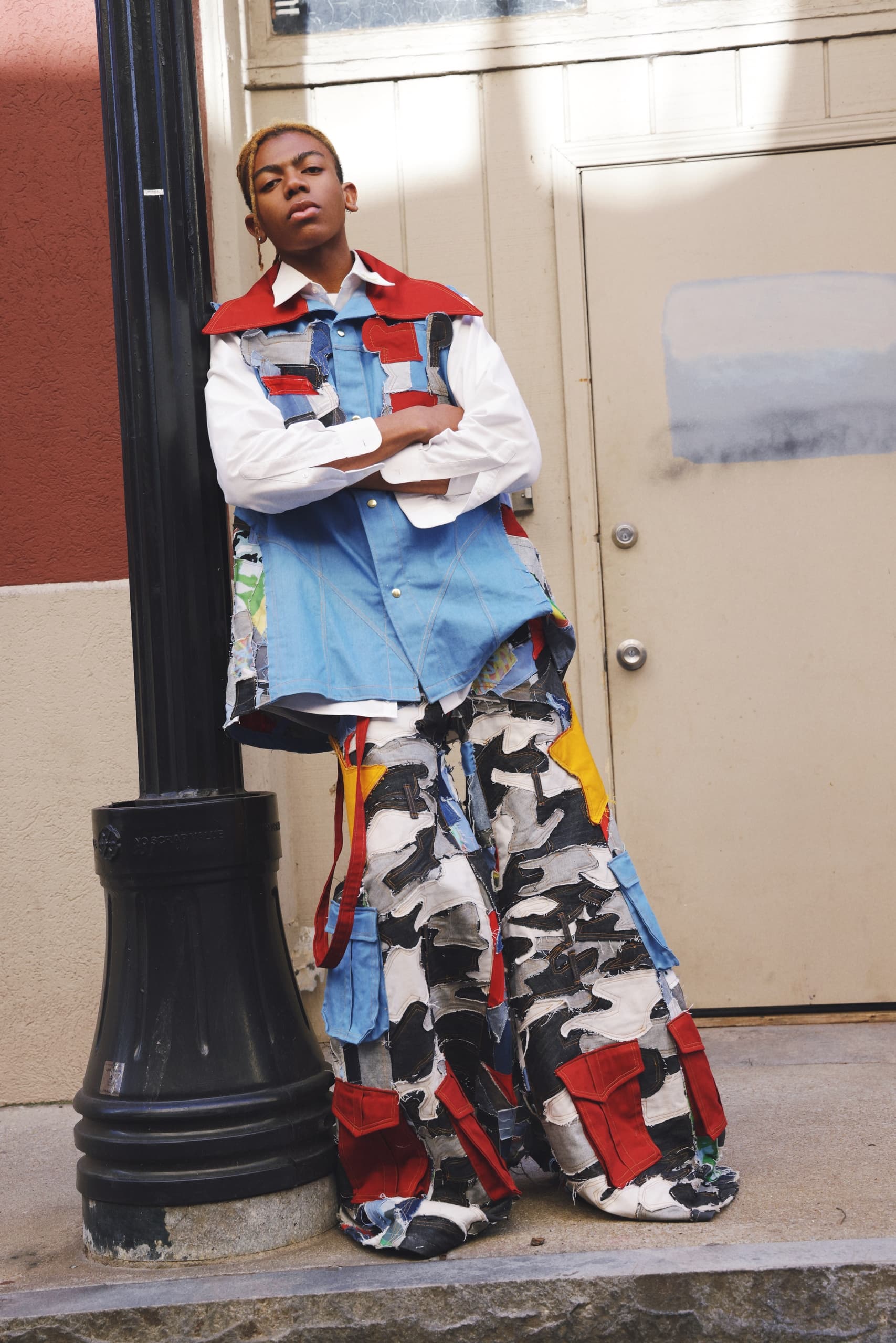 Person in colorful, artistic outfit leaning against a lamppost on urban street.