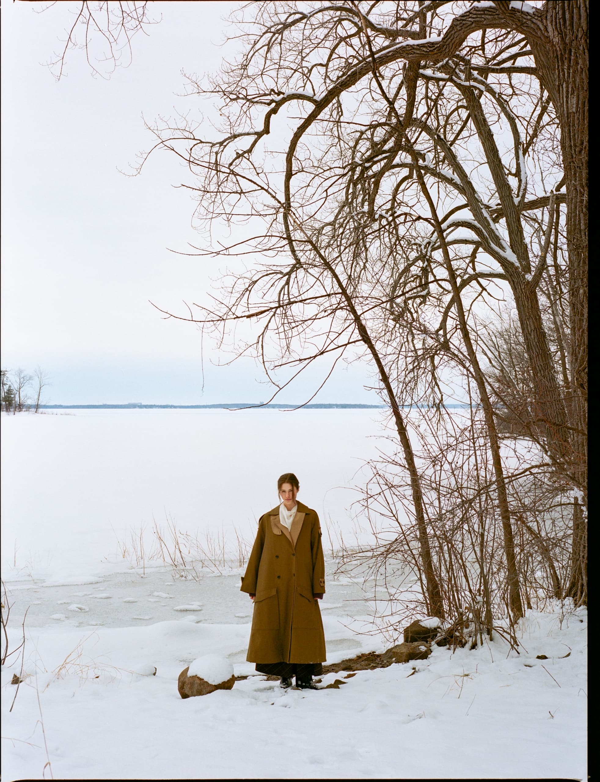 Person in long coat stands by a snowy, frozen lake with bare trees.