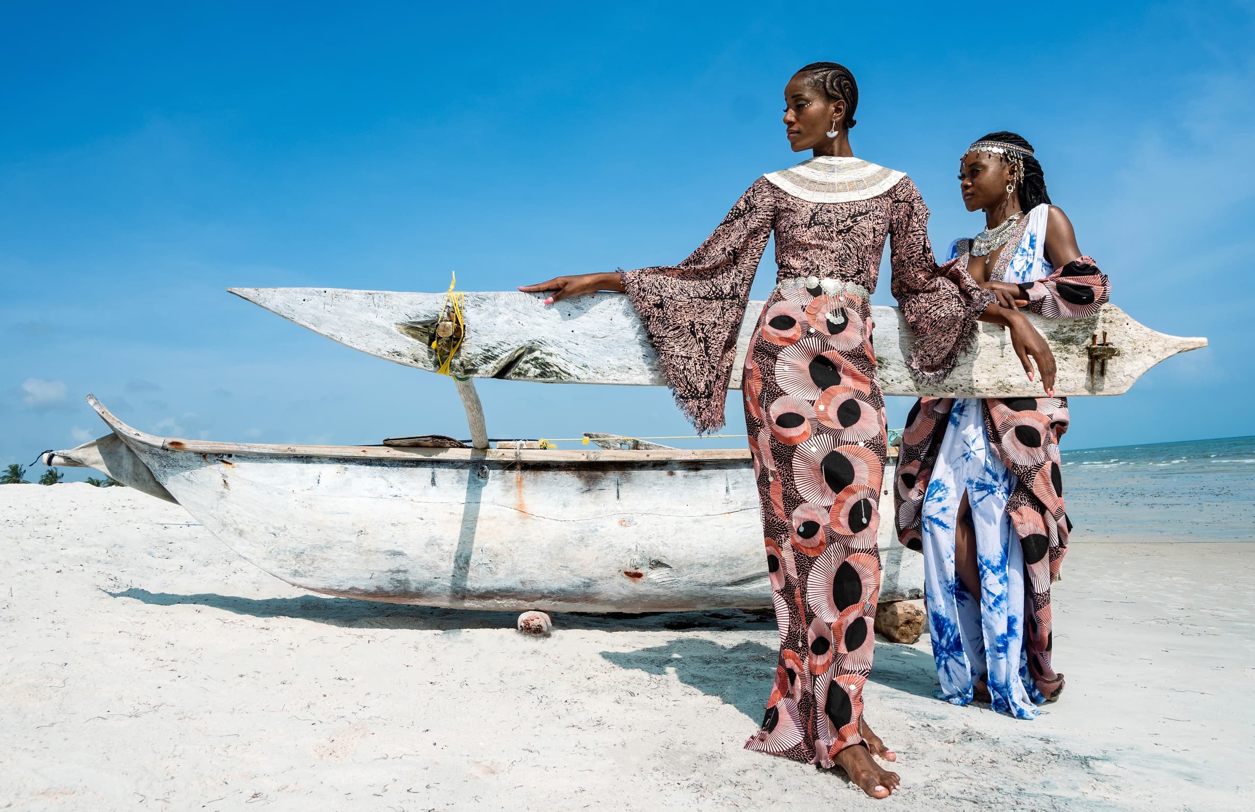 Fashionable women in vibrant dresses by a boat on a sunny beach.