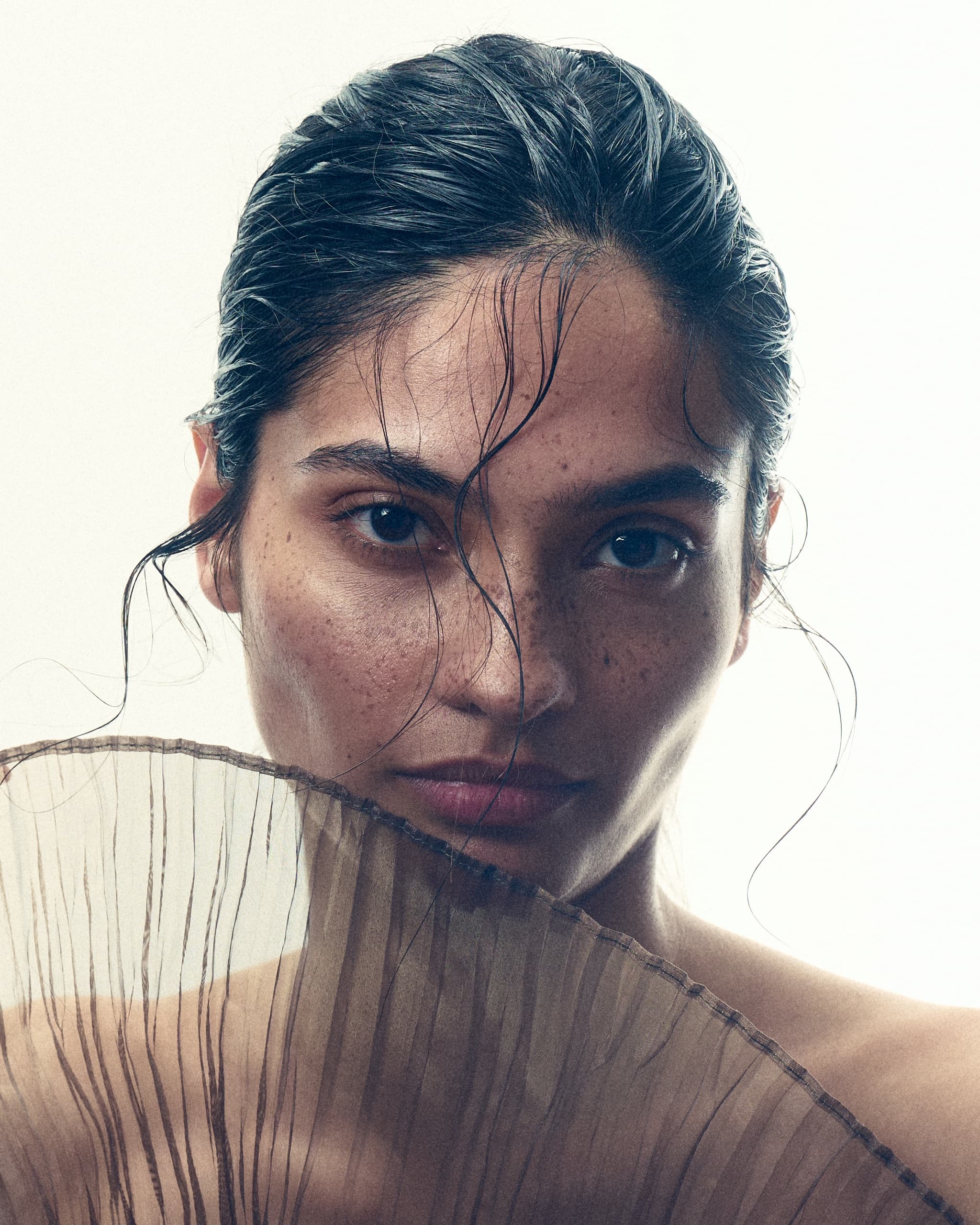 Portrait of a person with freckles holding a pleated fan, hair sleekly styled.
