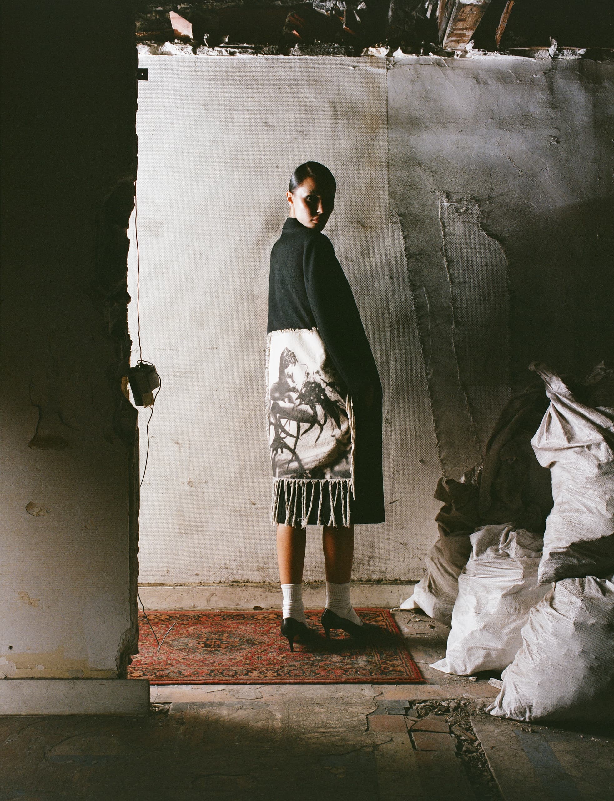 Fashion model in artistic coat standing in a dimly lit, rustic room.