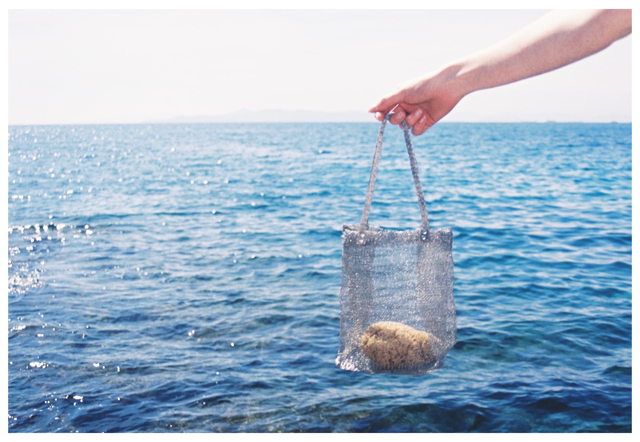 Hand holding a mesh bag with a sponge by the sea under a bright blue sky.