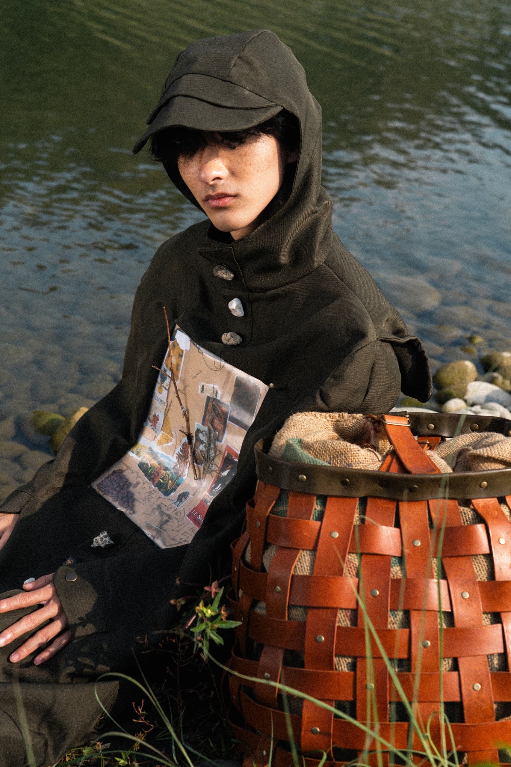 Person in hooded outfit sitting by a river with a woven basket.