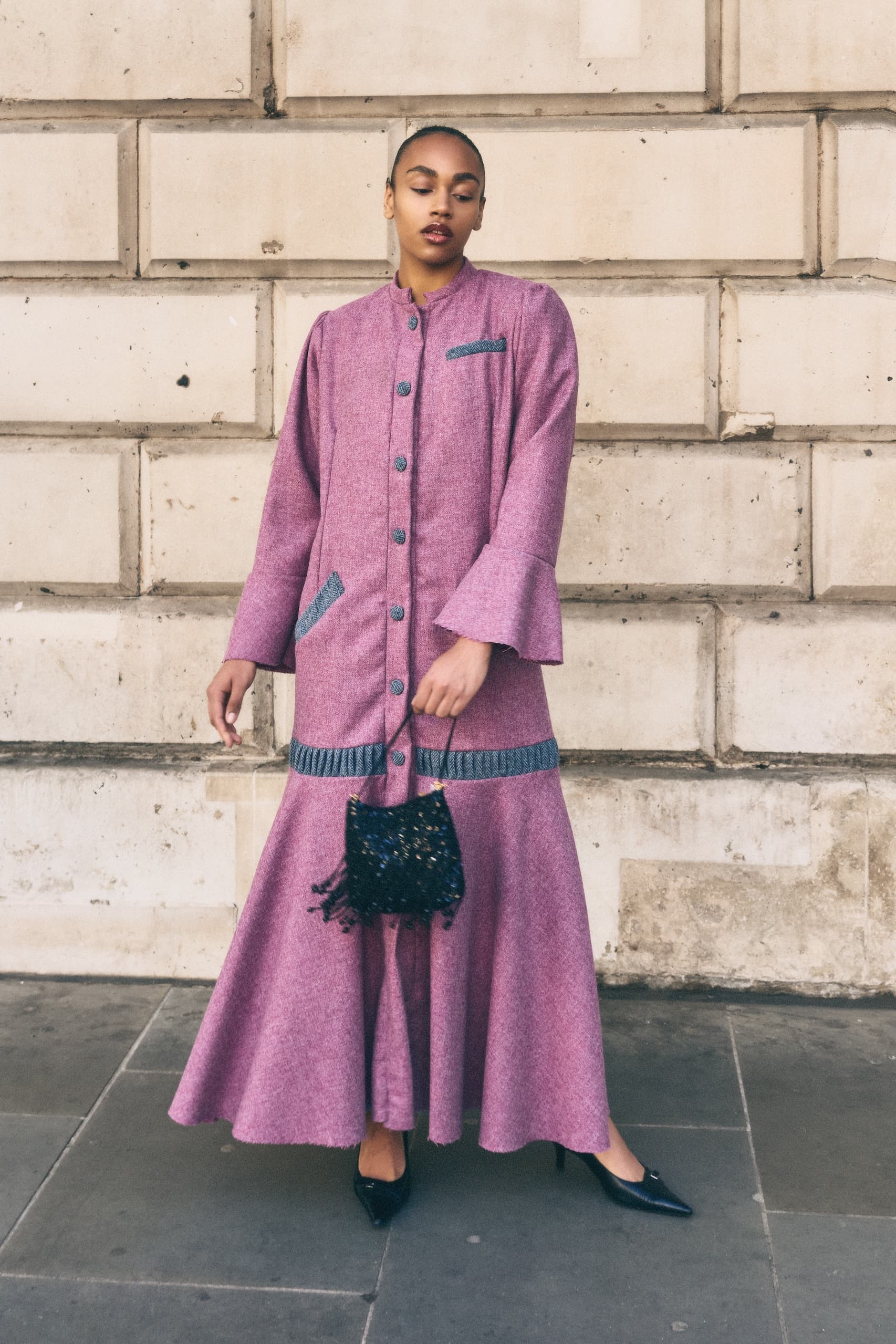 Woman in elegant pink coat dress with black purse, standing by a stone wall.