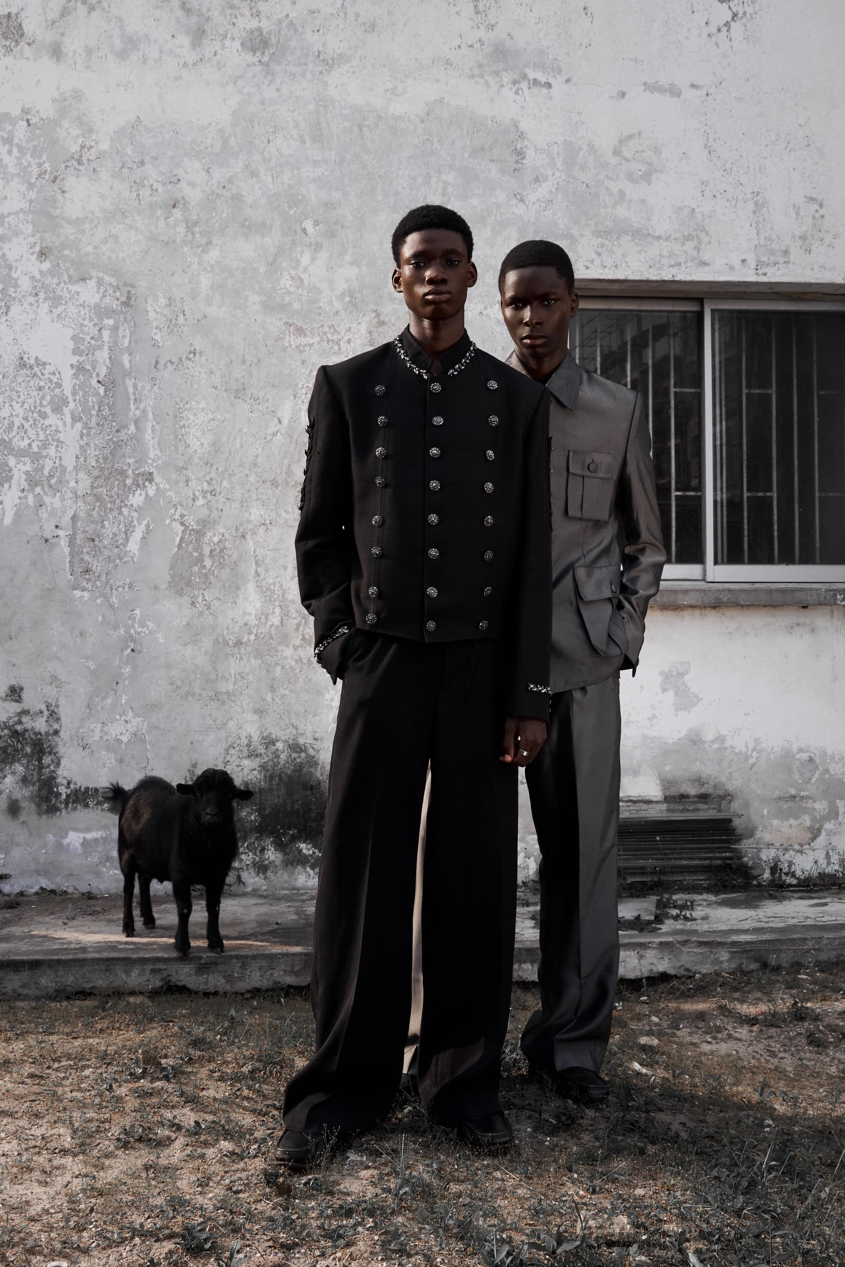 Two men in stylish clothing with a goat in an urban outdoor setting.
