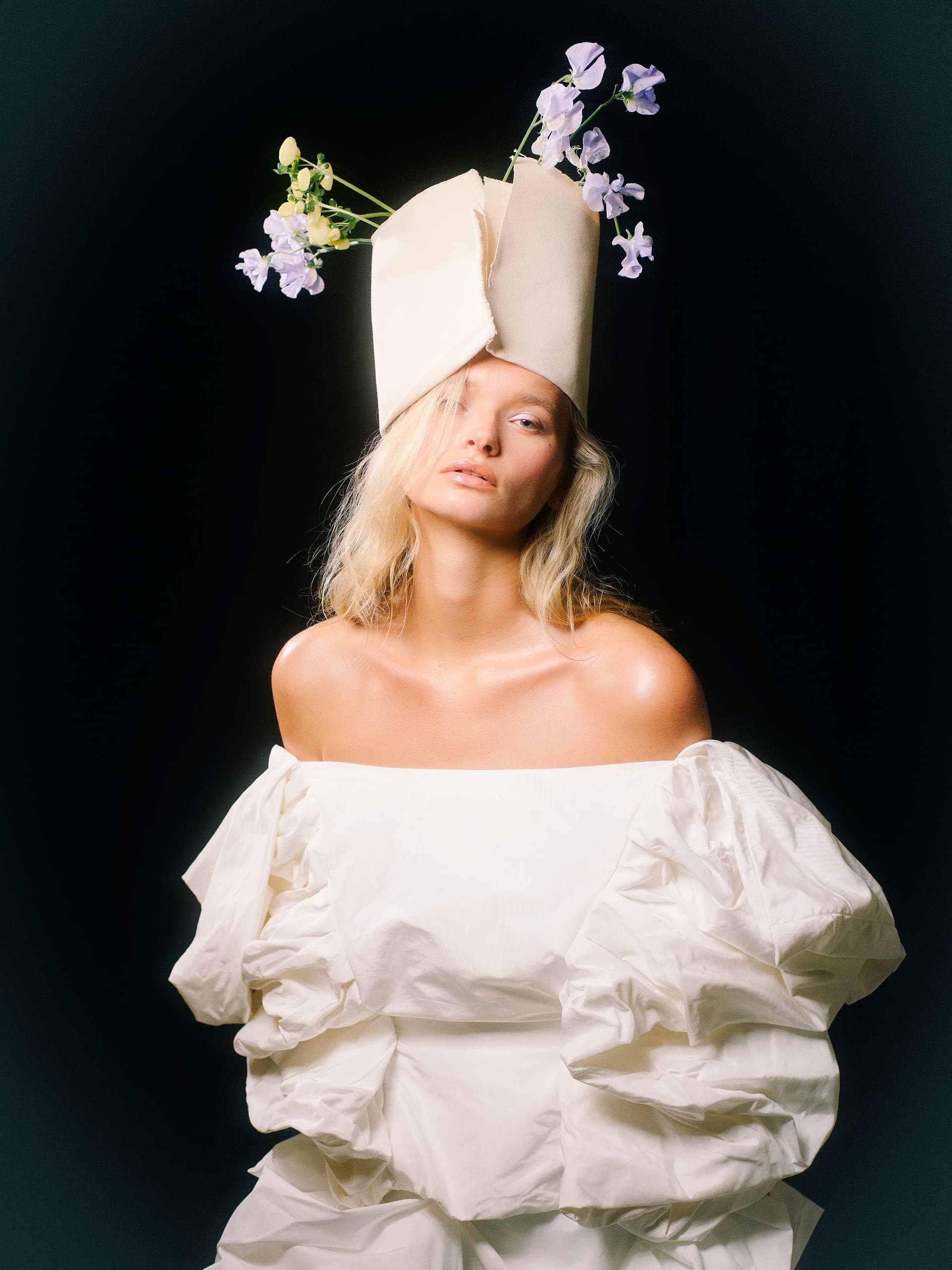 Blonde woman in a unique floral hat and white dress against a dark background.
