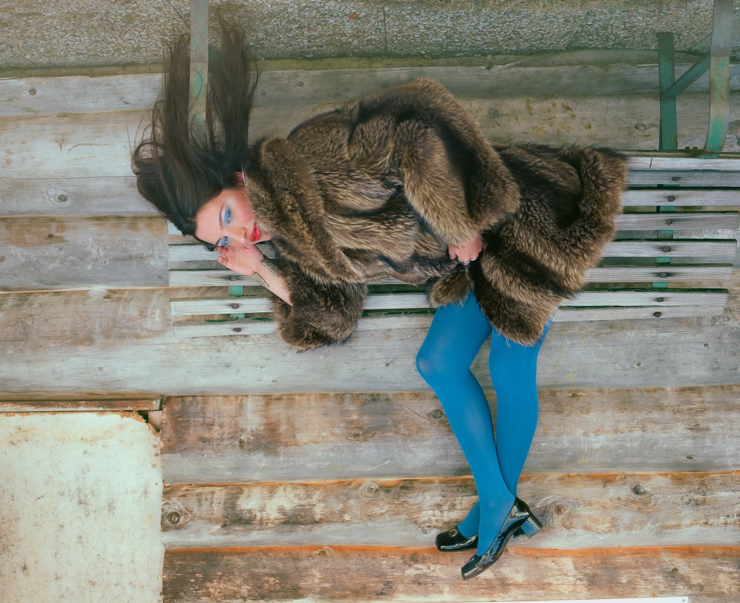 Woman in fur coat with bright blue tights lying on bench, rustic wooden background.