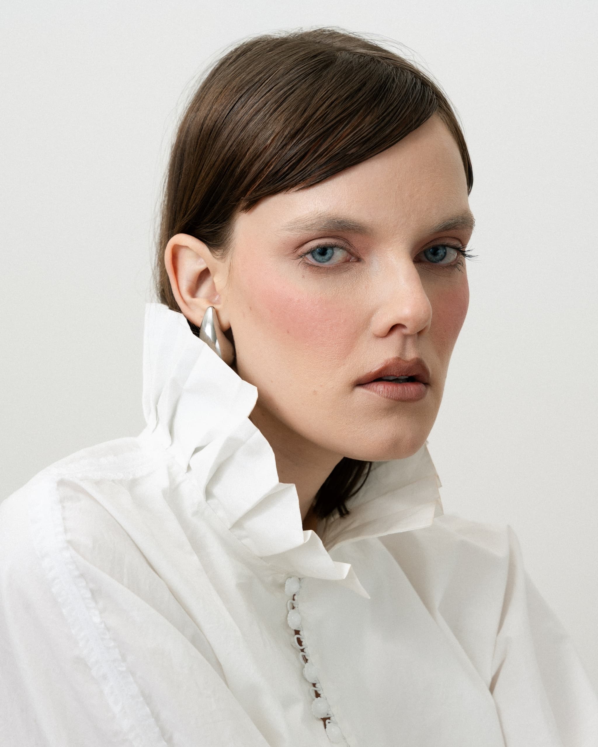 Close-up of a model in a ruffled white blouse, with sleek brown hair and sharp blue eyes.