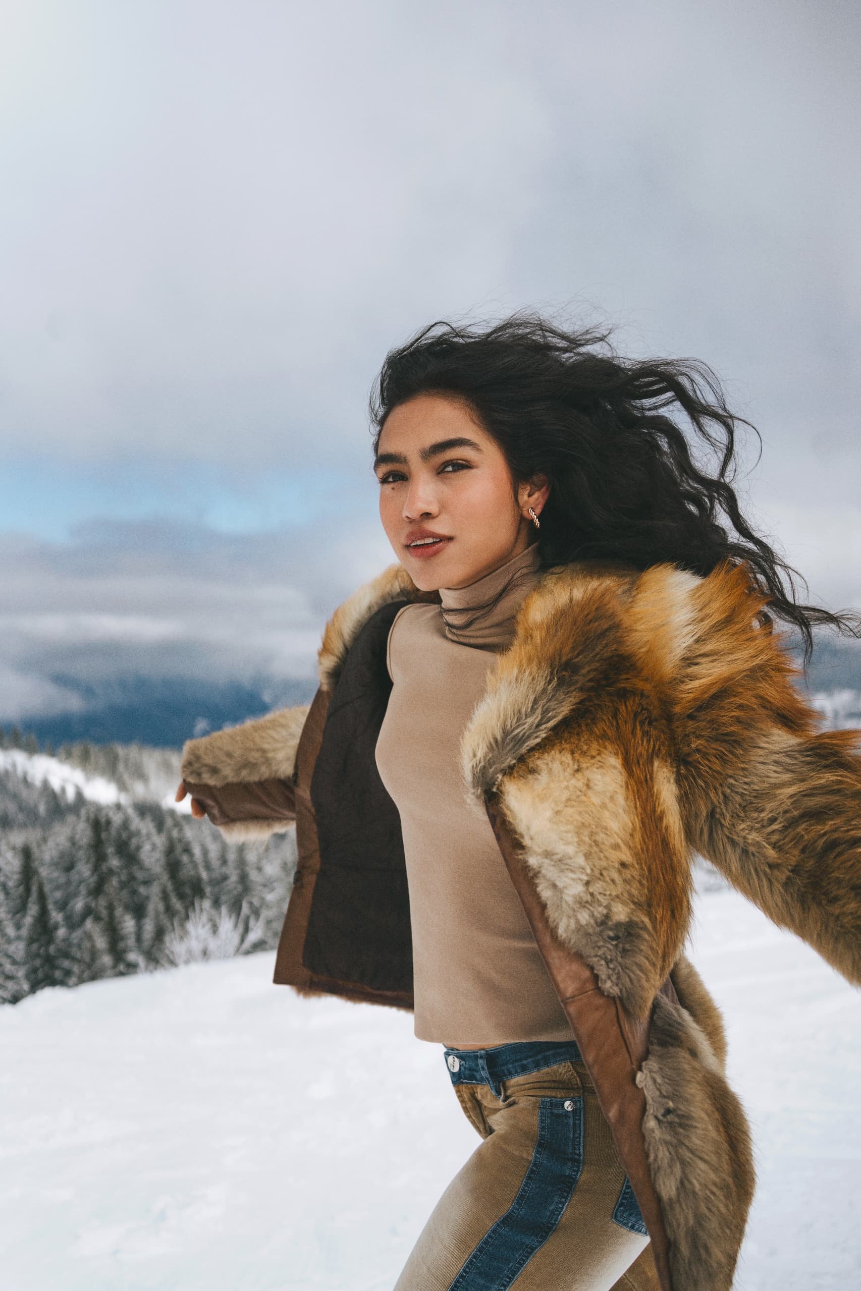 Woman in fur coat enjoying snowy mountain landscape with flowing hair and confident smile.
