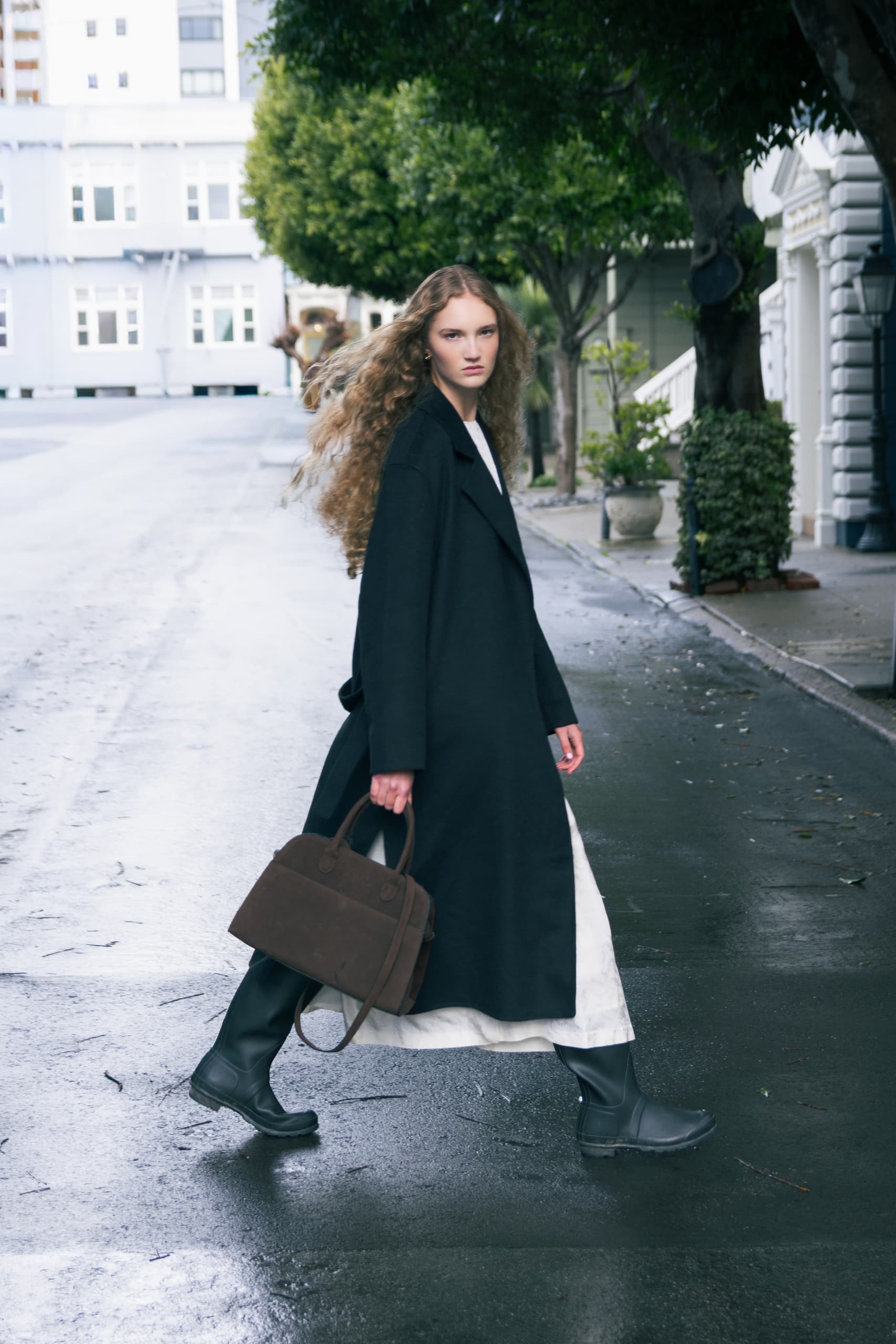 Woman in a black coat walking on a rainy street, holding a brown bag.