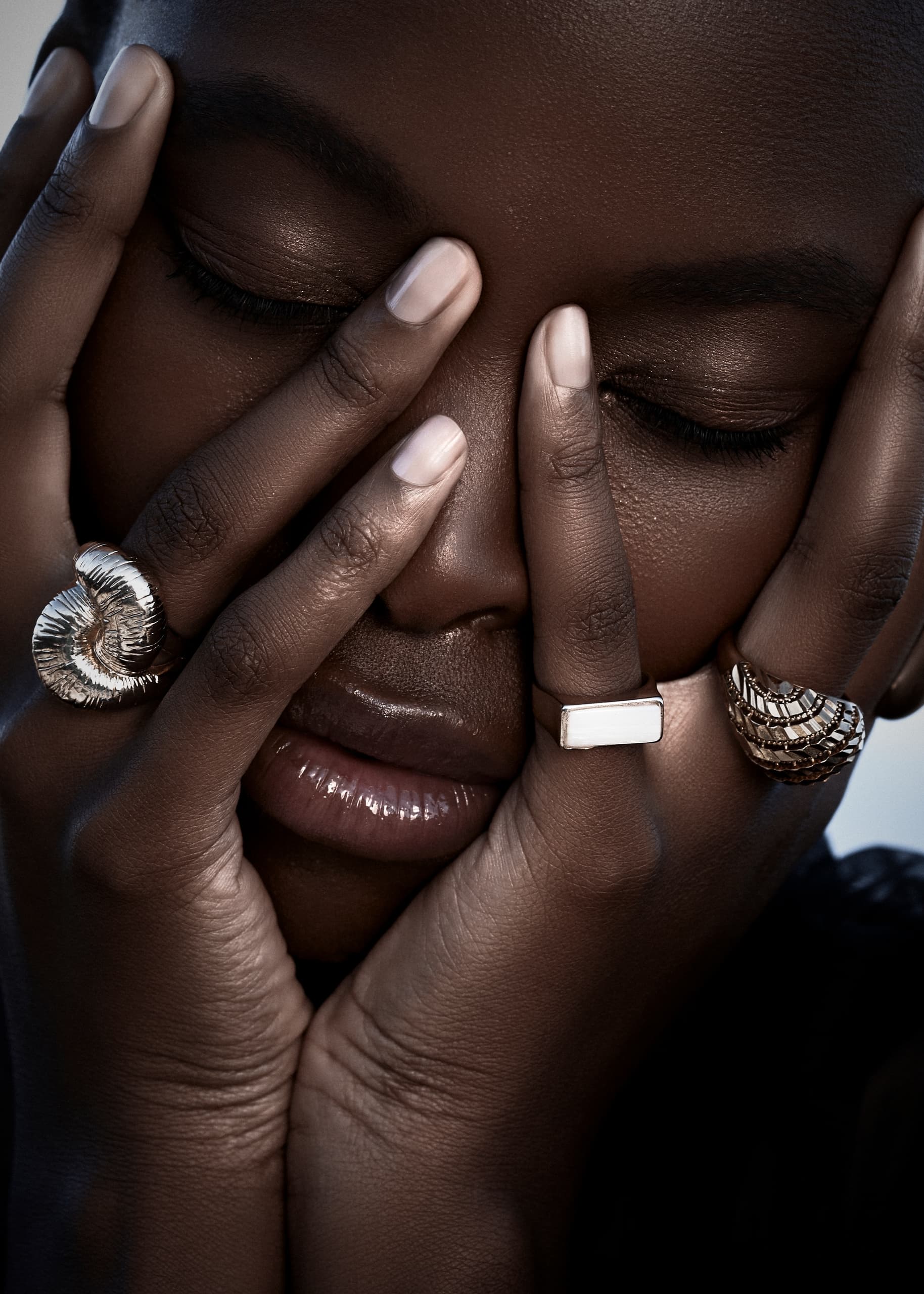 Close-up of hands with stylish rings covering a serene face.