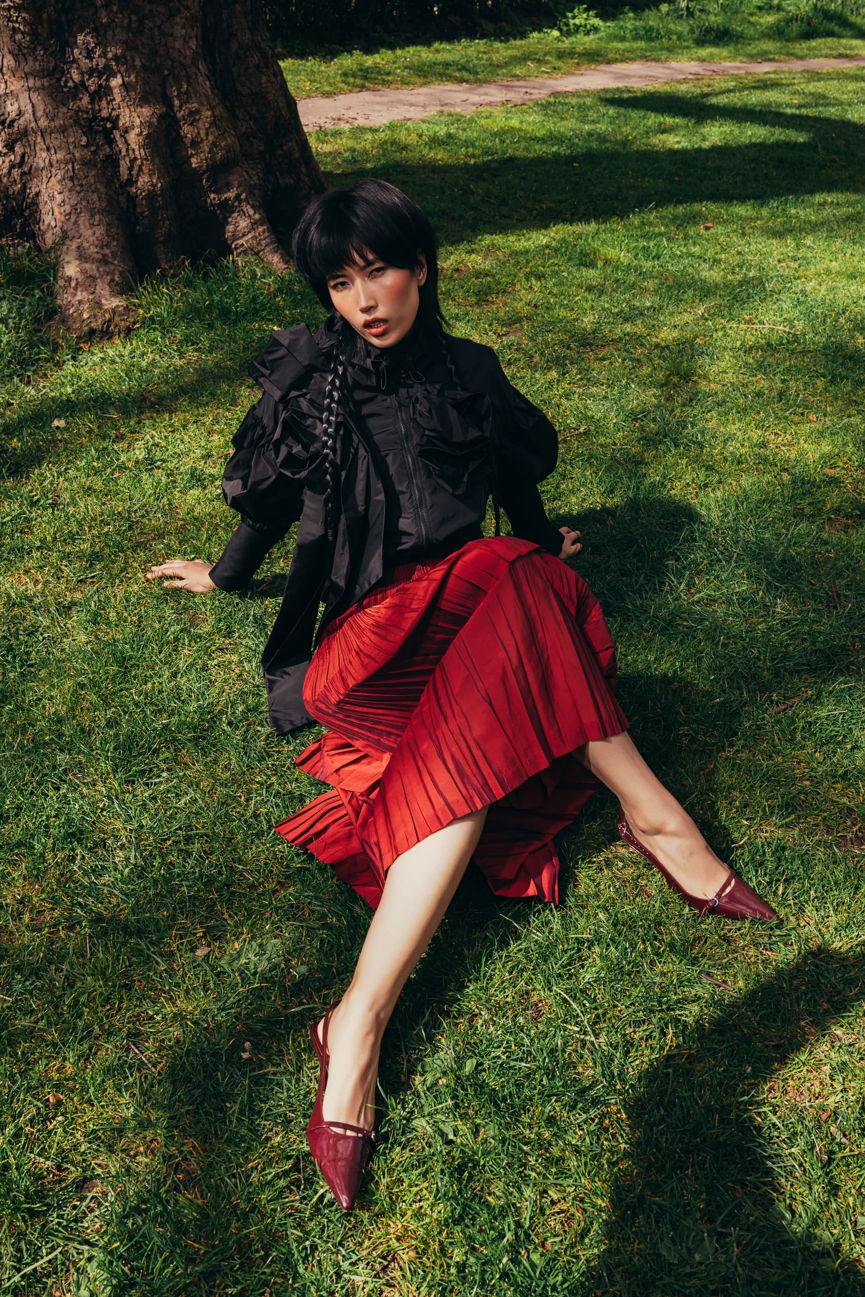 Woman in black blouse and red skirt sitting on grass by a tree.