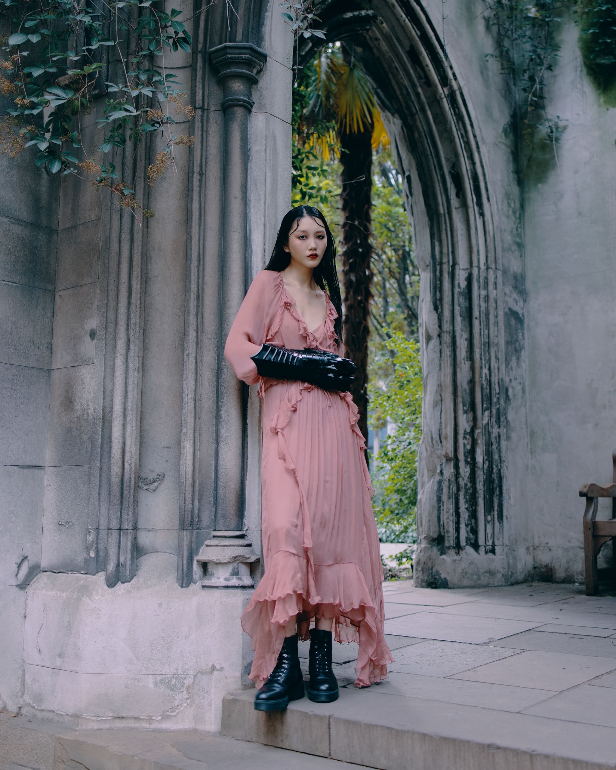 Woman in a flowing pink dress and black boots in a gothic stone archway.