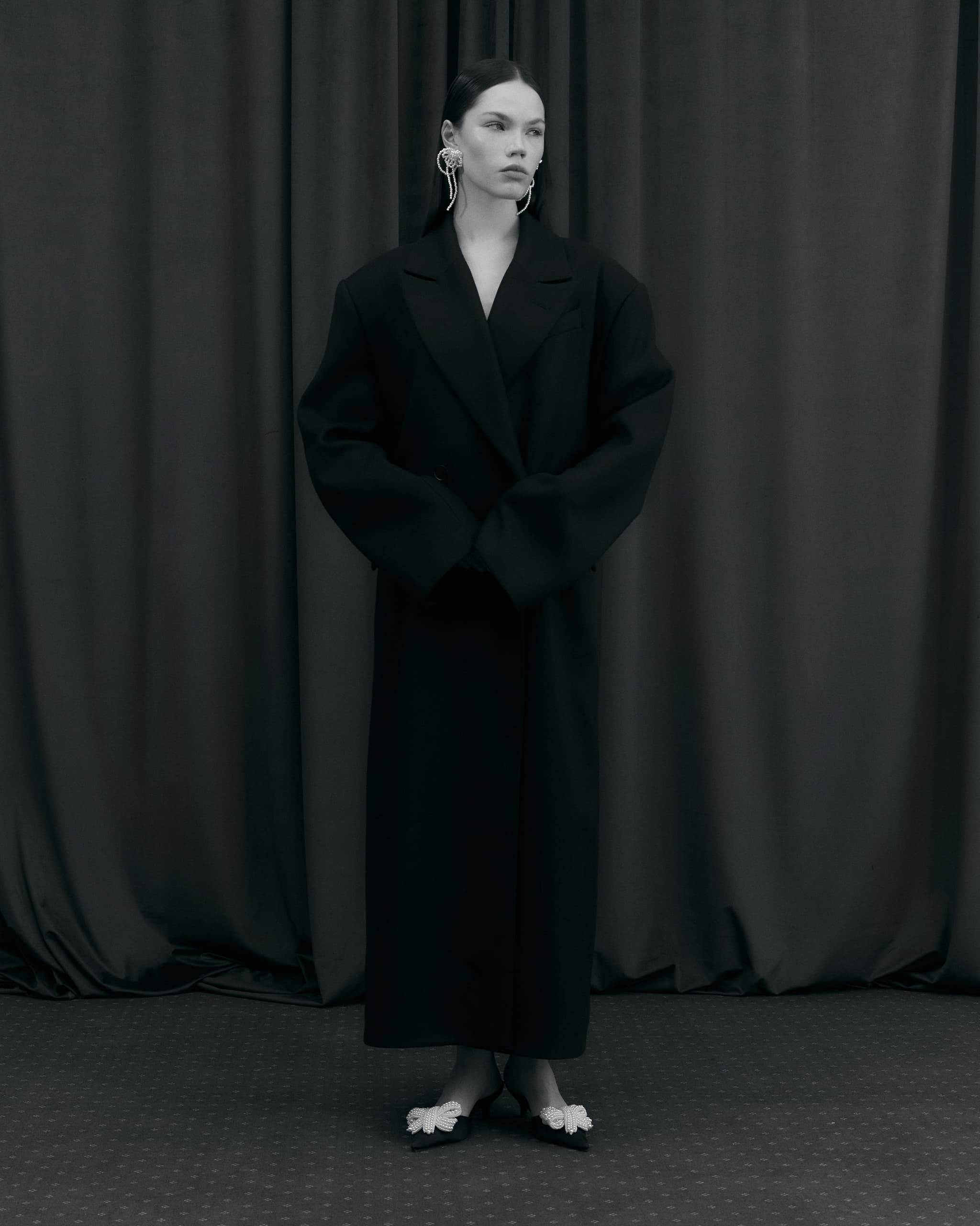 Person in oversized black coat with earrings, standing against dark curtains.