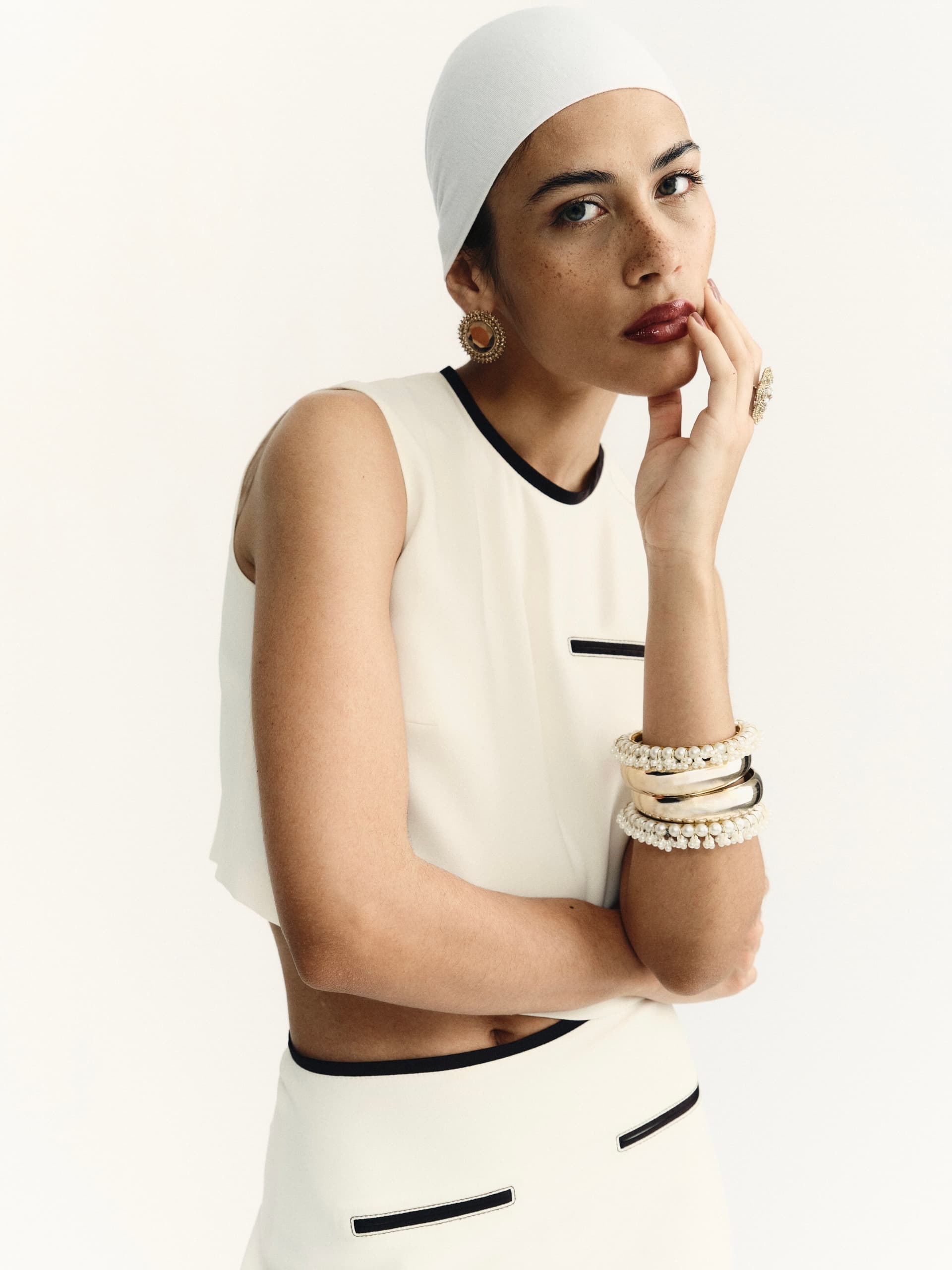Fashionable woman in white outfit with elegant jewelry and headscarf, posing thoughtfully.