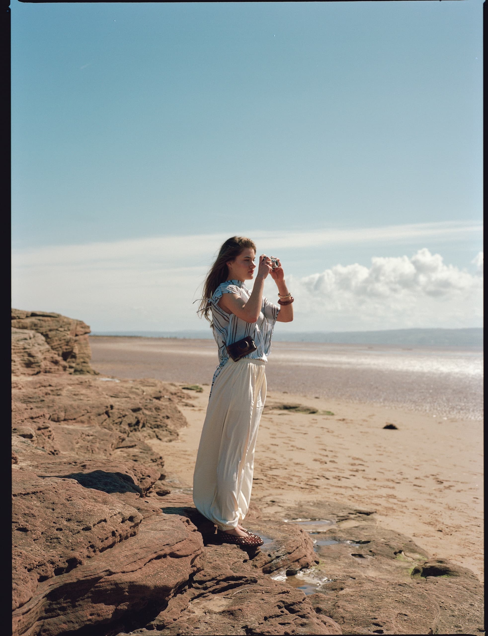 Woman photographing beach scenery on a sunny day, standing on rocks by the sea.