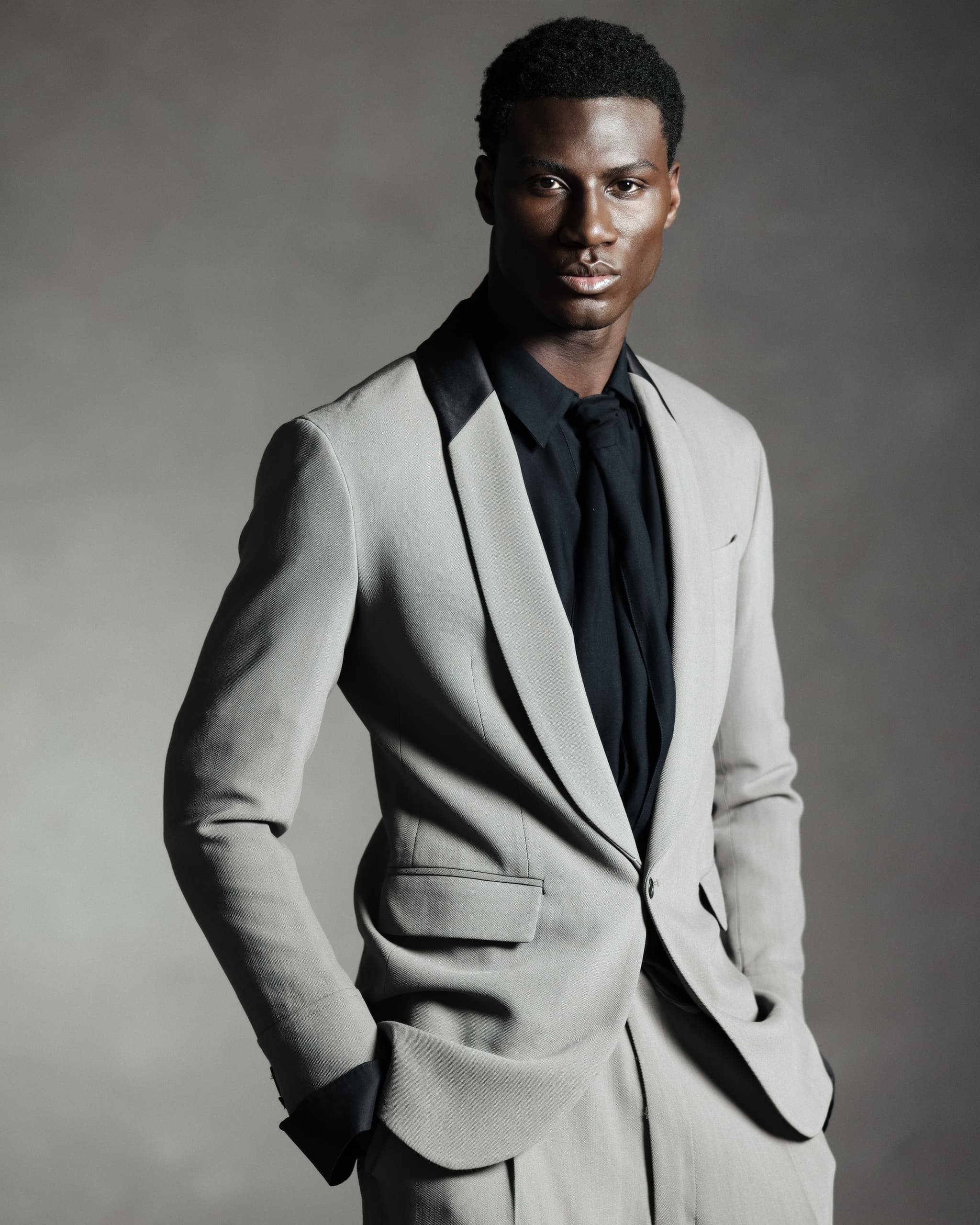 Man in stylish beige suit with black shirt and tie, hands in pockets.