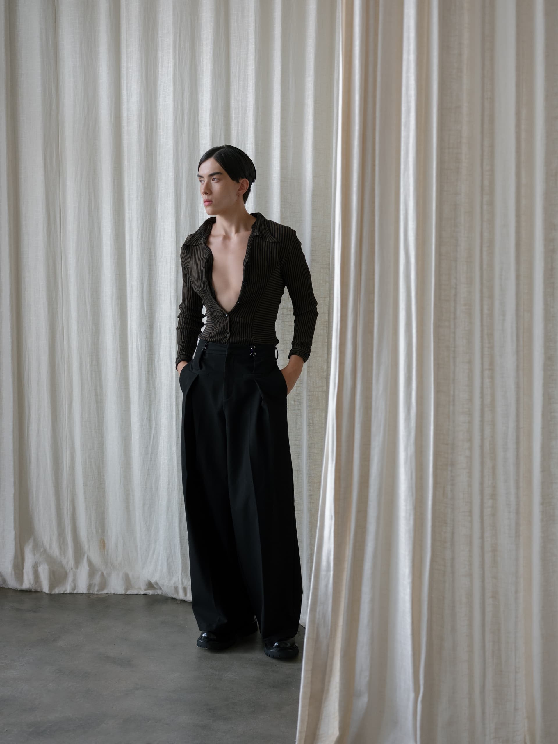 Person in stylish black outfit by white curtains, striking a thoughtful pose.