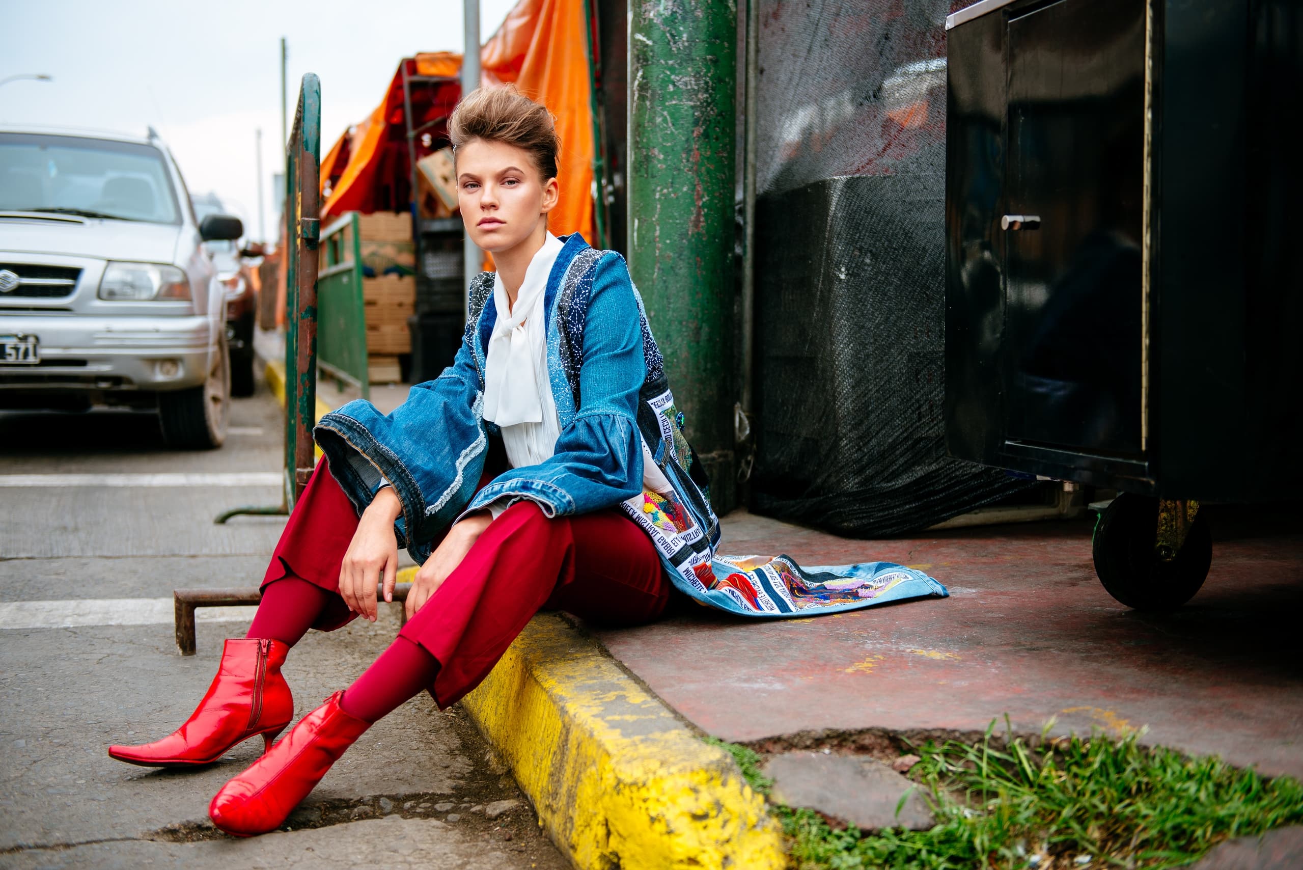 Fashionable person sits on curb in vibrant outfit and red boots on city street.