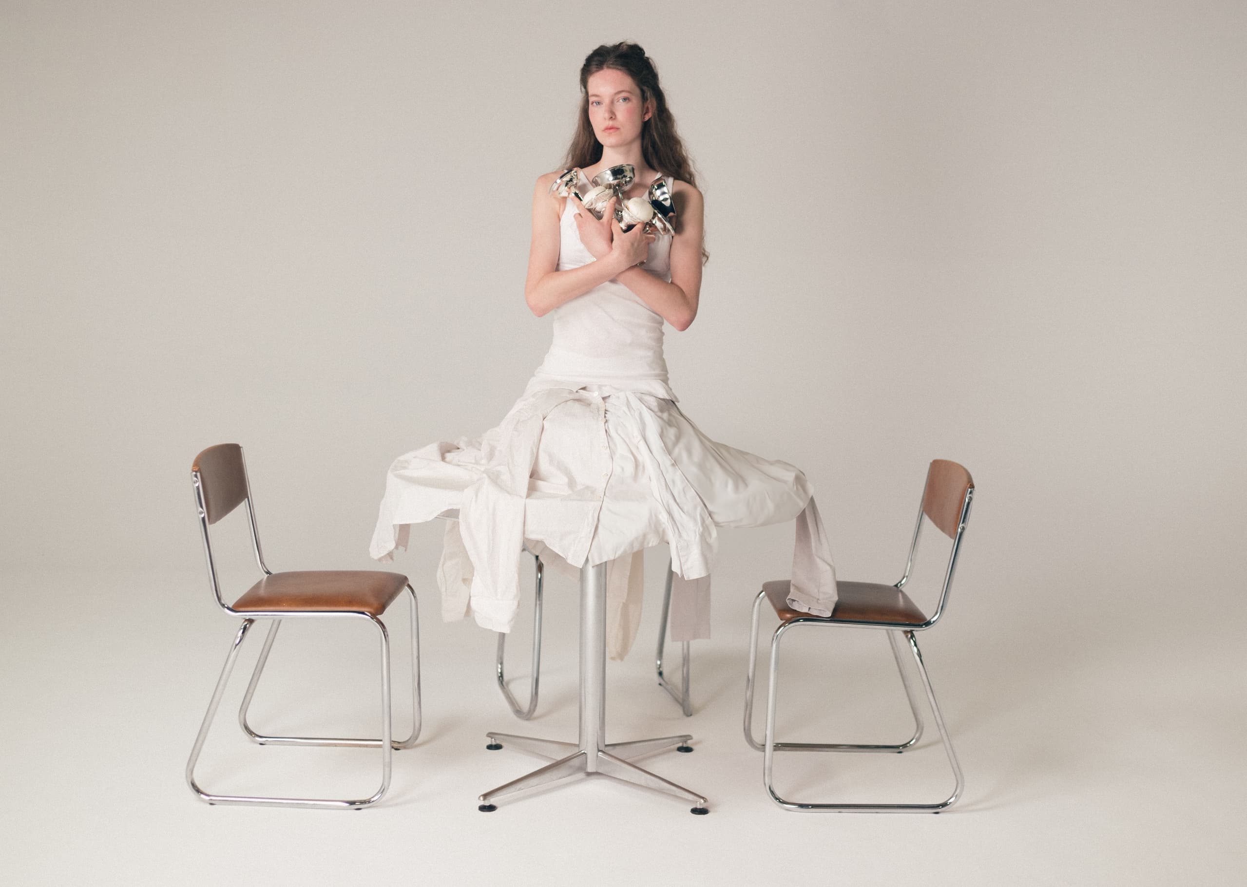 Woman in white dress sitting on table between chairs, holding chrome objects.