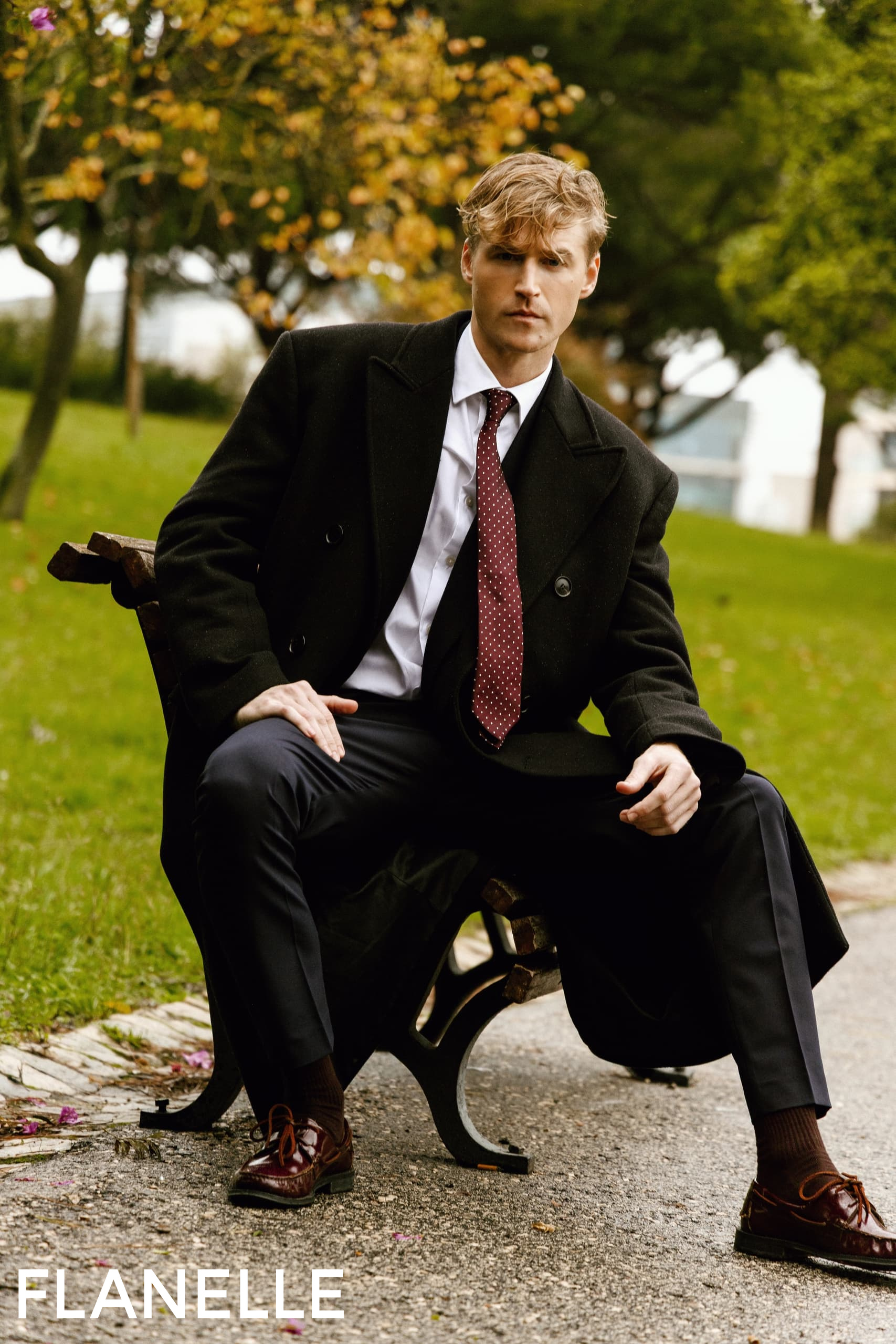 Man in formal wear sitting on a park bench, wearing a black coat and red tie.