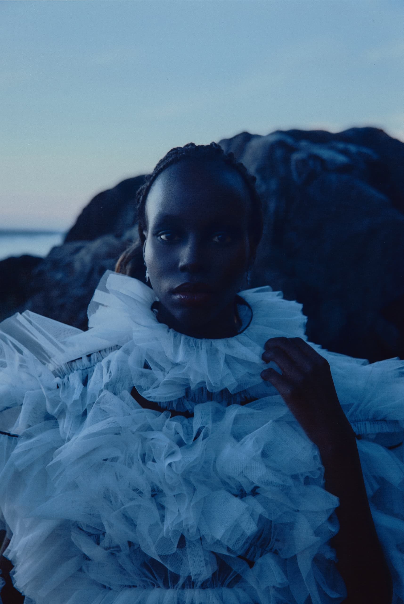Person in elegant ruffled dress at twilight by rocky landscape and sea.