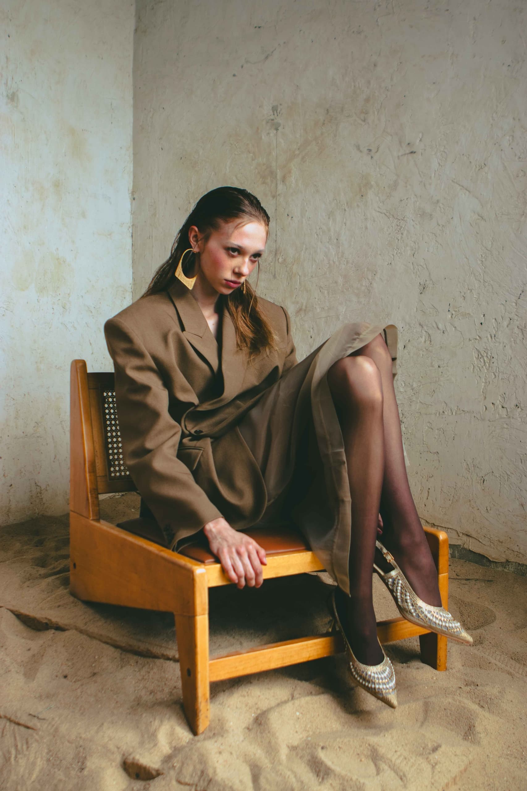 Woman in oversized blazer sitting on a wooden chair in a minimalist setting.